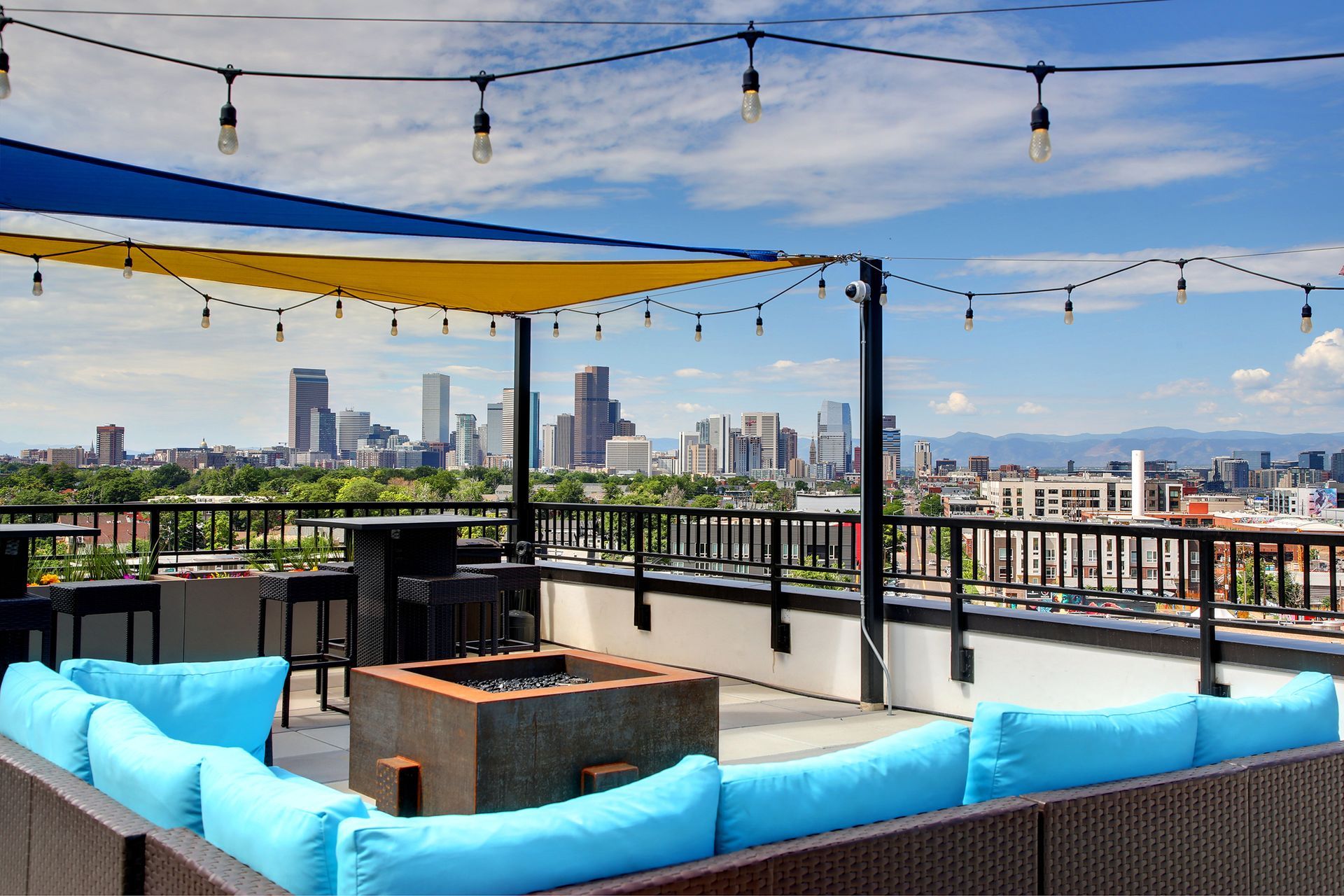 Rooftop patio with cityscape view, blue cushions, fire pit, and string lights under a blue and yellow awning.