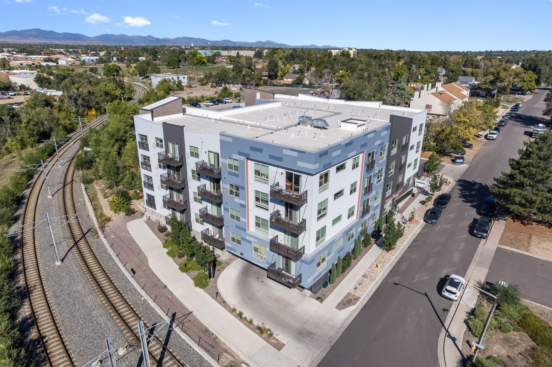 Aerial view of a modern apartment building with blue and gray siding next to railroad tracks and a street with cars.