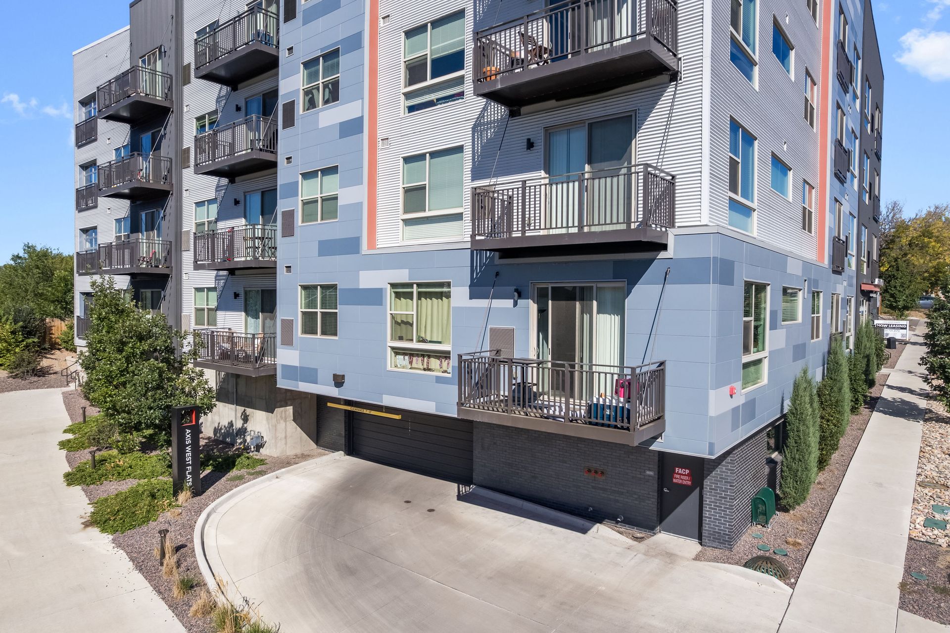 Modern apartment building with balconies, gray and blue facade, driveway entrance.