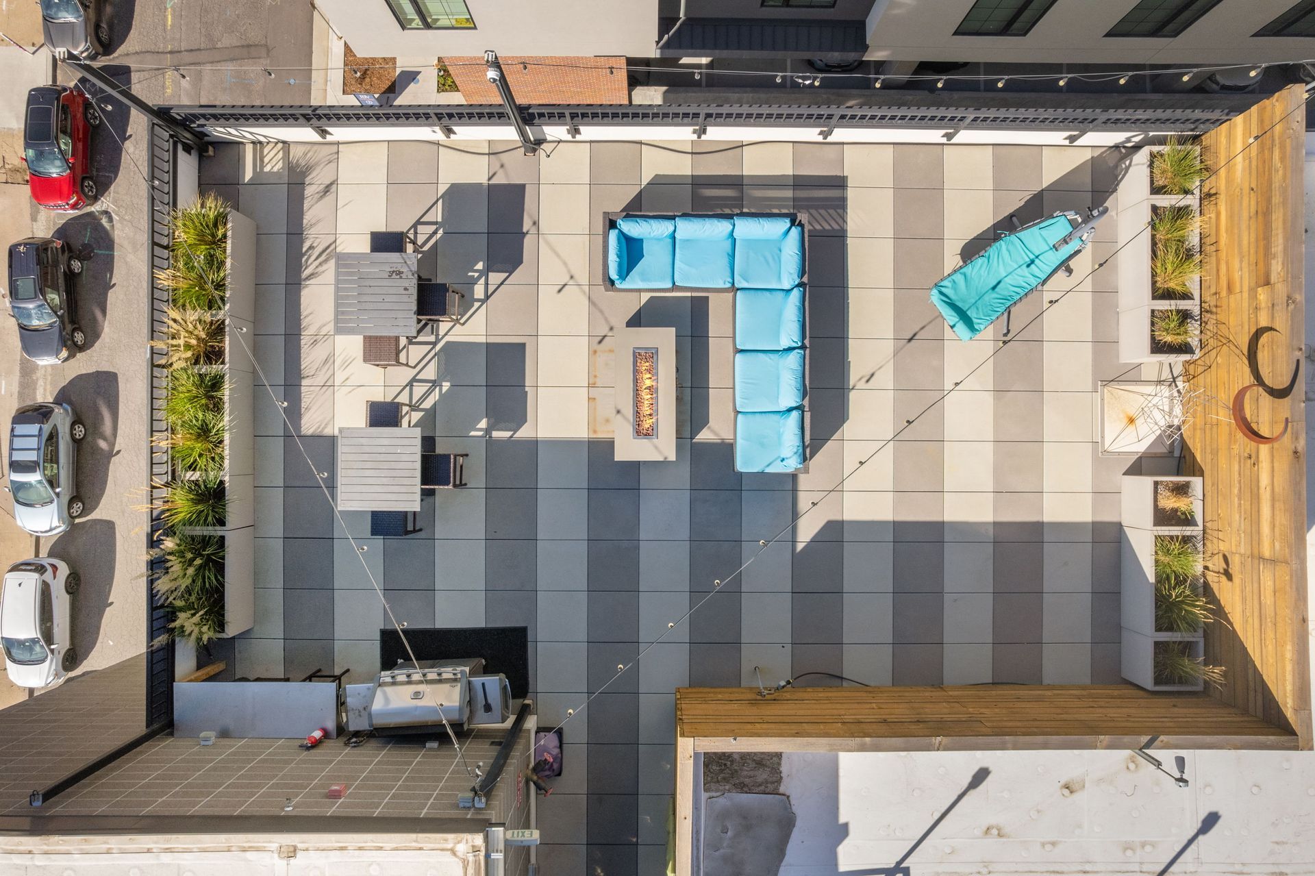 Overhead view of a rooftop patio with seating, fire pit, and string lights, surrounded by buildings and cars.