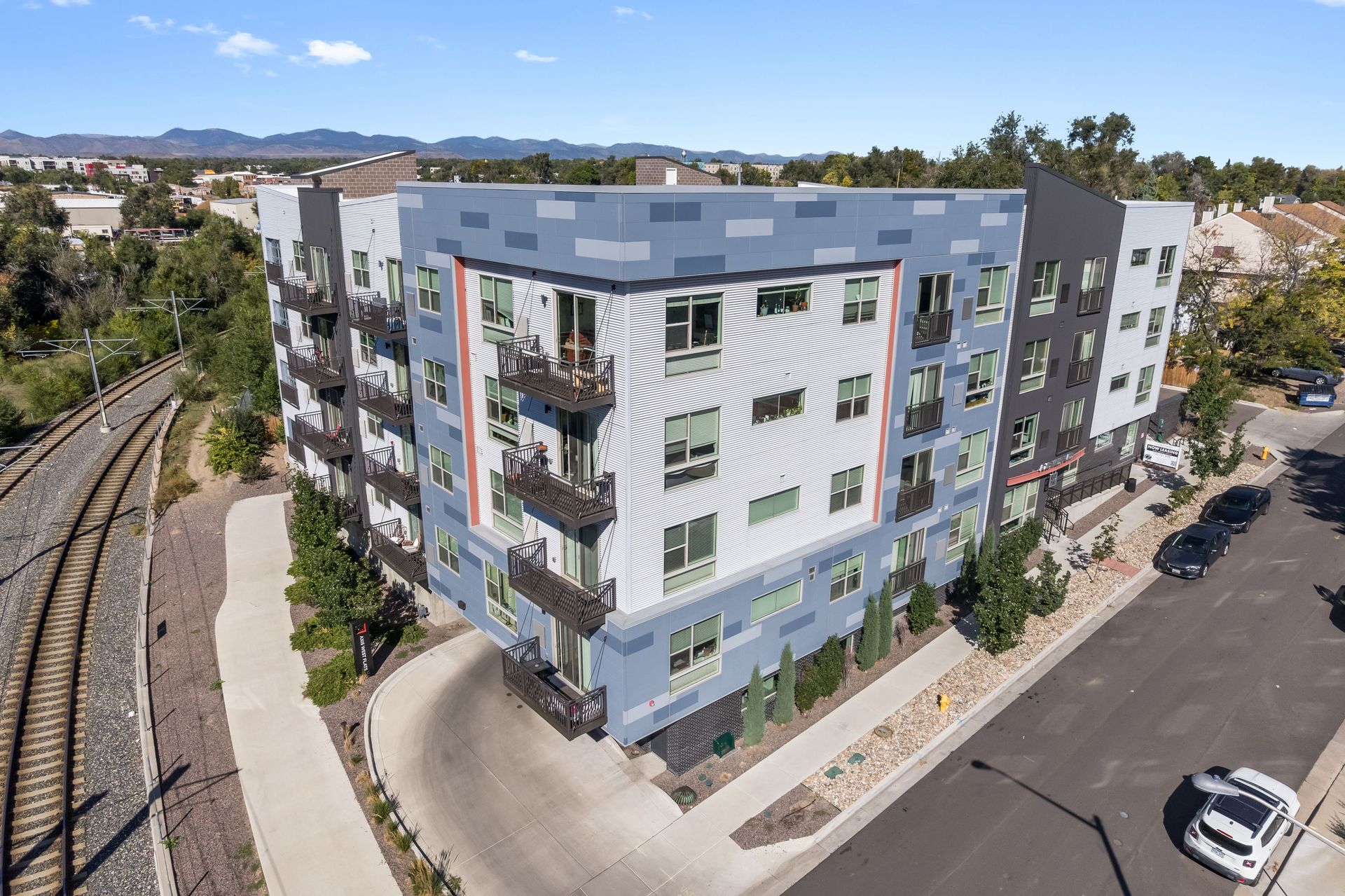 Multi-story apartment building with blue, gray, and orange accents, near railroad tracks and a street.