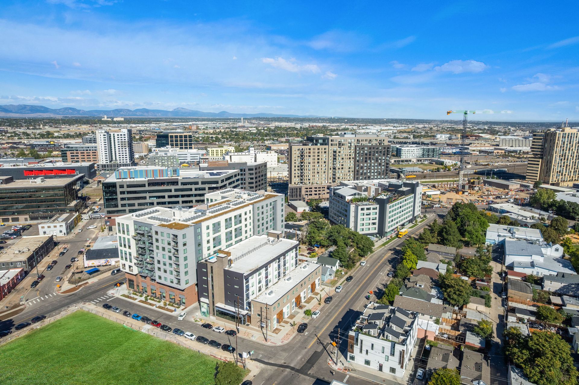 Aerial view of Denver cityscape with modern buildings under a blue sky, including a green field on the left.