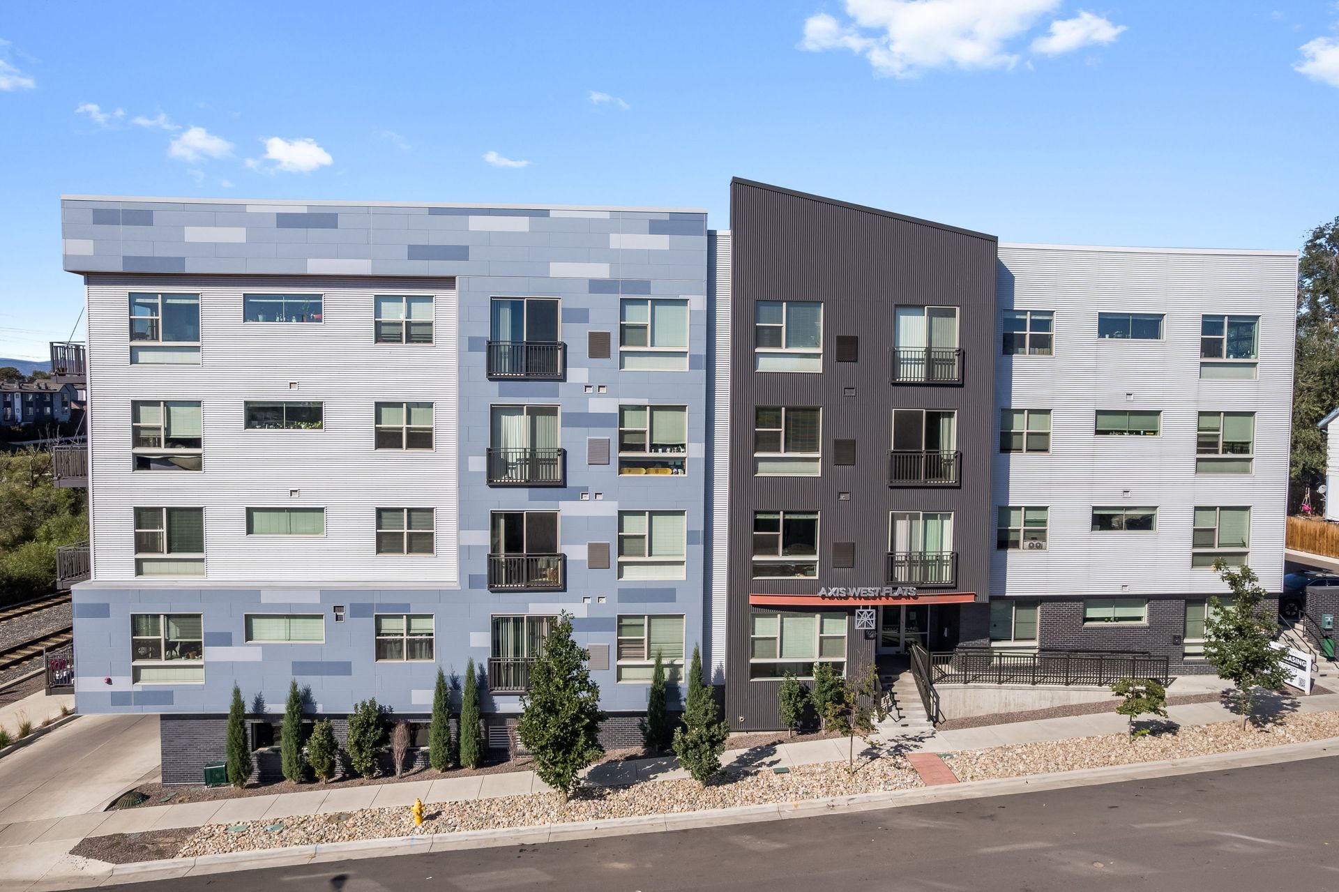 Modern apartment building with blue, gray, and black facade. Small balconies, trees, and a paved walkway in front.