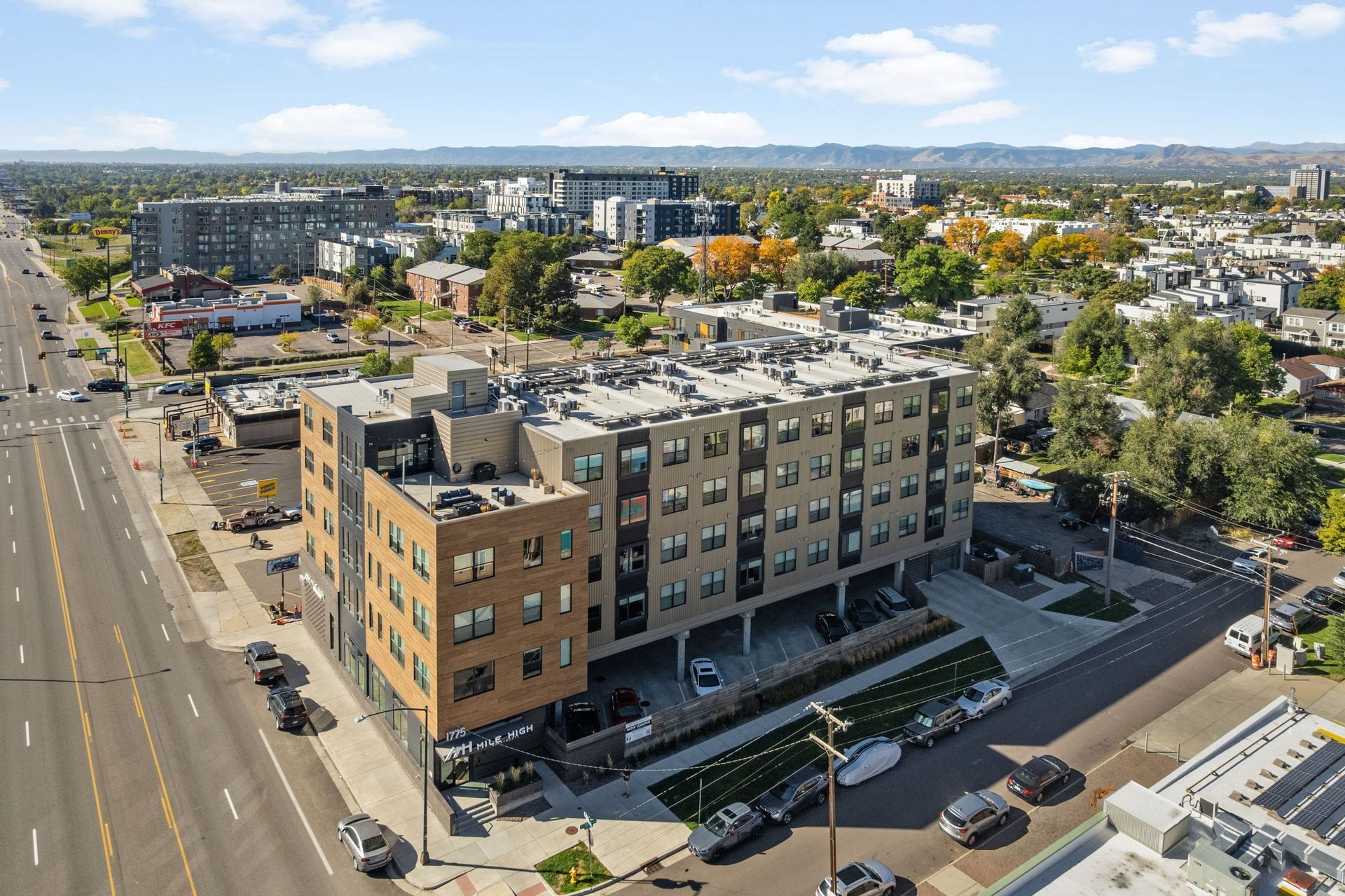 Multi-story apartment building with cars parked along a city street; trees, and other buildings visible in the background.
