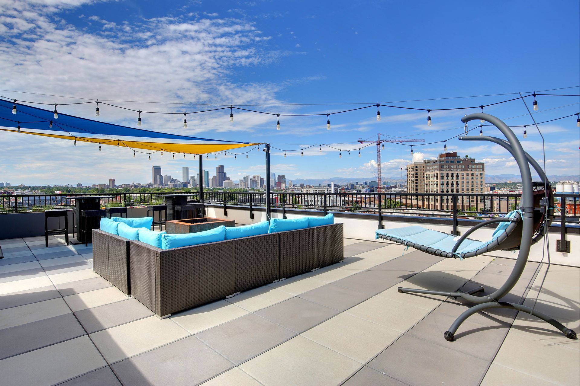 Rooftop lounge with cityscape view, blue cushions, a hanging chair, string lights, and a blue and yellow awning.