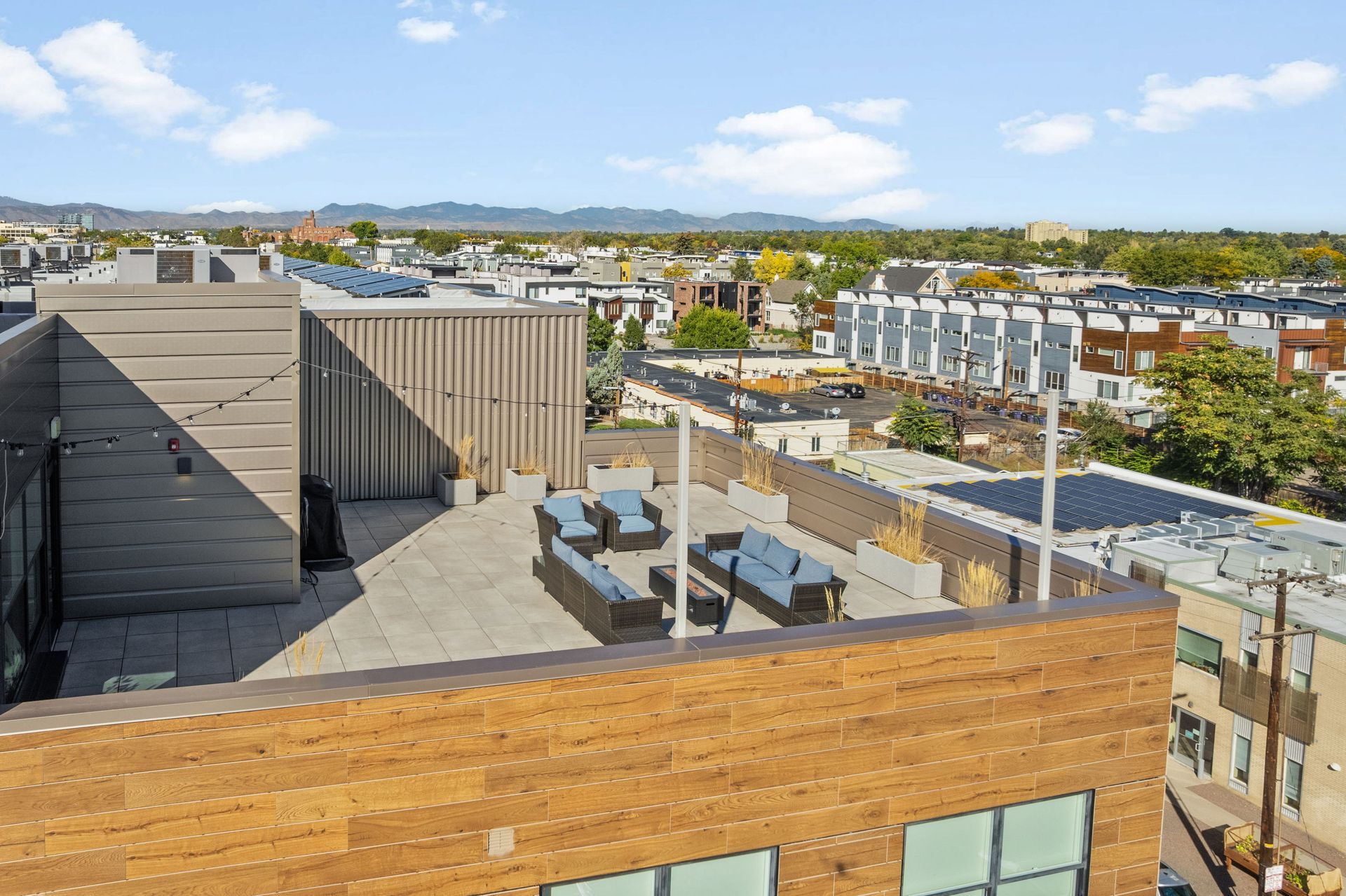 Rooftop patio with seating, wooden exterior. Cityscape and mountains in the background under a blue sky.