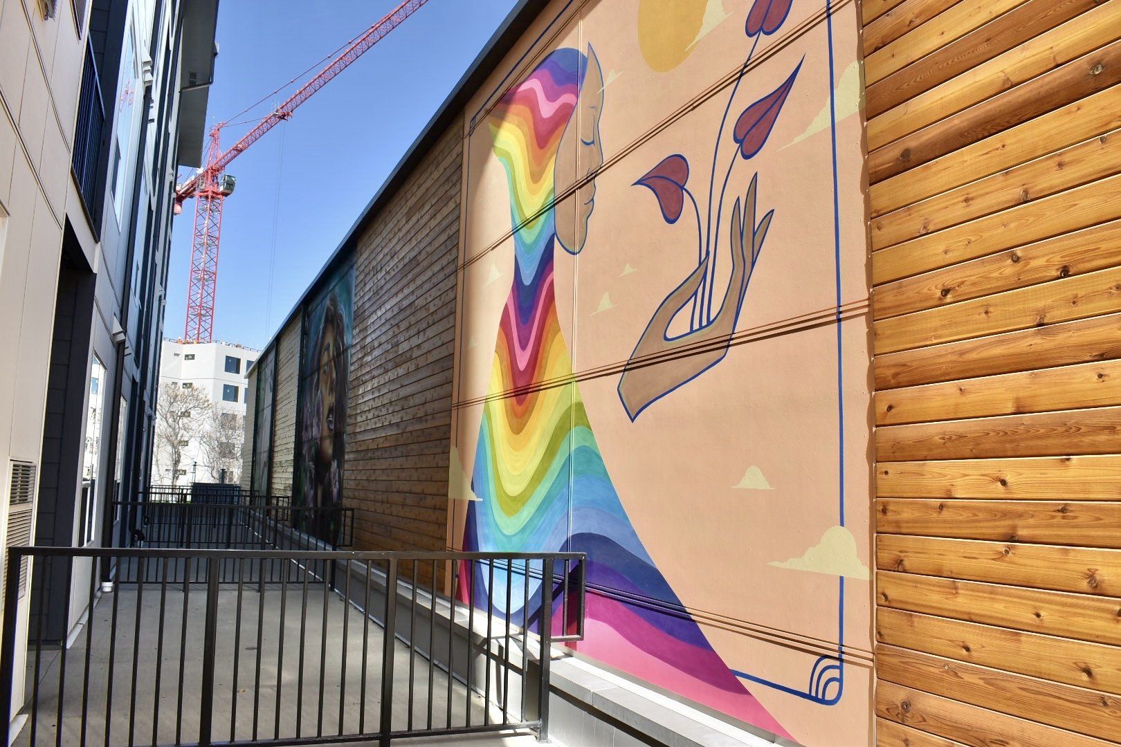 Mural of a figure with rainbow dress, holding leaves, next to wooden wall. Construction crane in background.