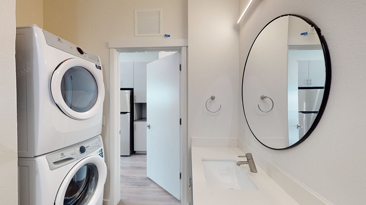 Stacked white washer and dryer next to a bathroom with a sink, round mirror, and an open doorway to a kitchen.