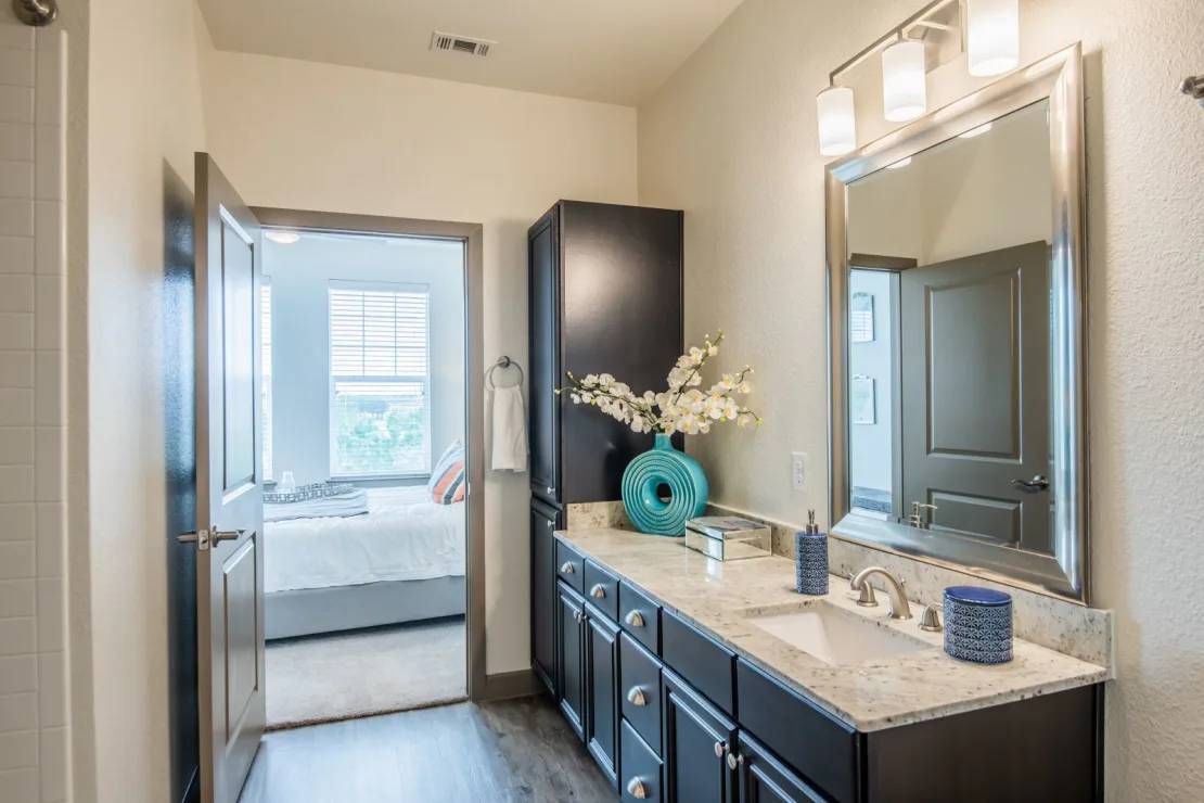 Modern bathroom with dark vanity, large mirror, and doorway to a bedroom with a bed by the window