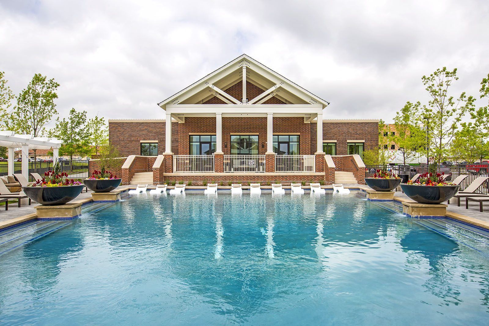Outdoor resort pool with a brick clubhouse and white gabled roof, flanked by potted plants and lounge chairs