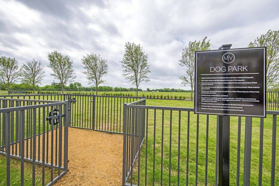 Black metal gate by a path in a grassy field, with a sign and trees under cloudy sky