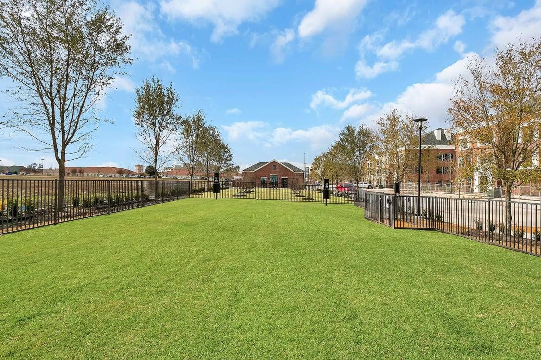 Grassy park with a fenced path, trees, and apartment buildings under a blue sky.