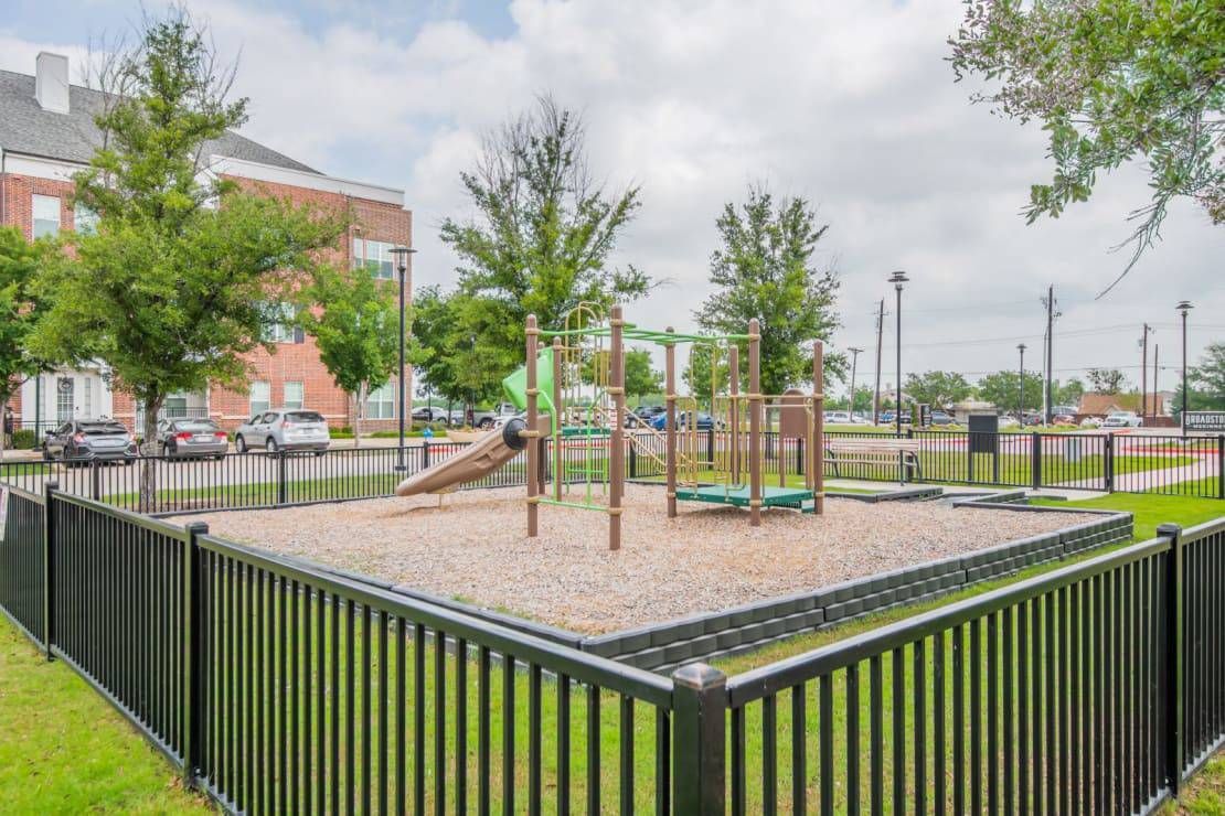 Fenced playground with slides and climbing structures in a park beside apartment buildings
