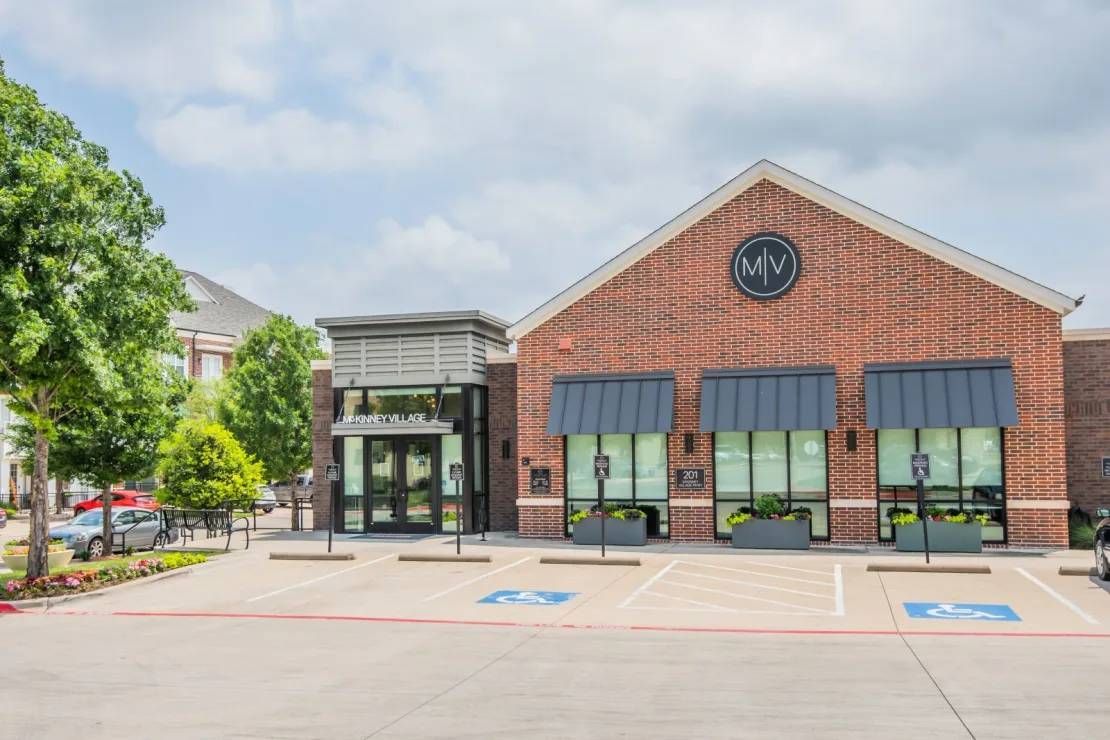 Brick storefront with black awnings and glass entrance, viewed from a parking lot on a sunny day
