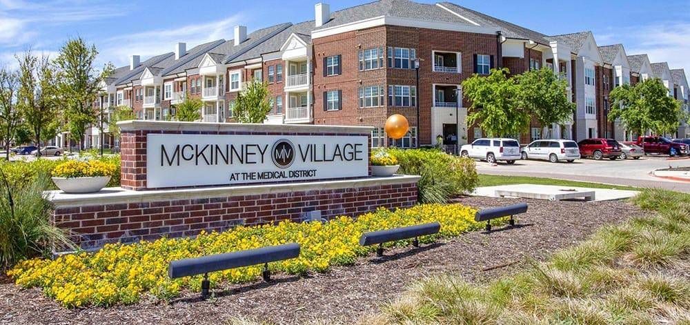 McKinney Village sign in front of brick apartment buildings and landscaped flower beds