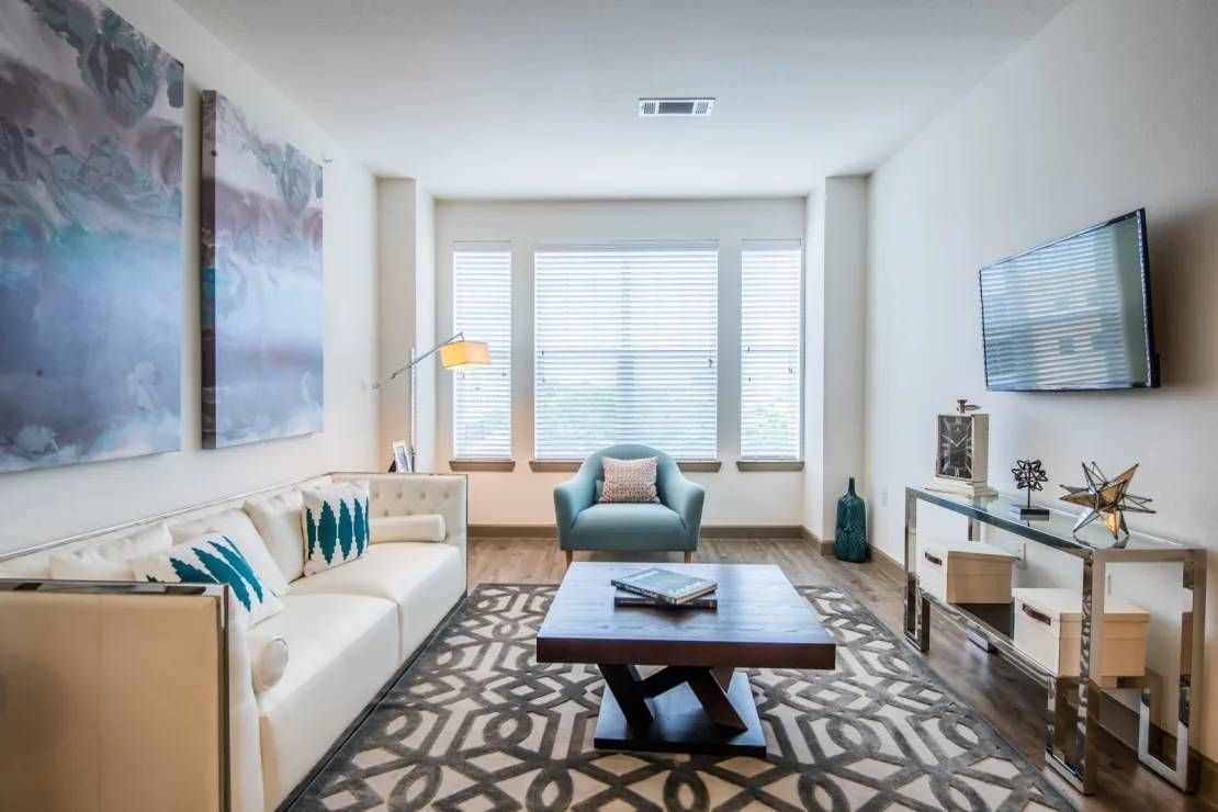 Bright modern living room with white sofa, teal chair, patterned rug, wall art, and large window.