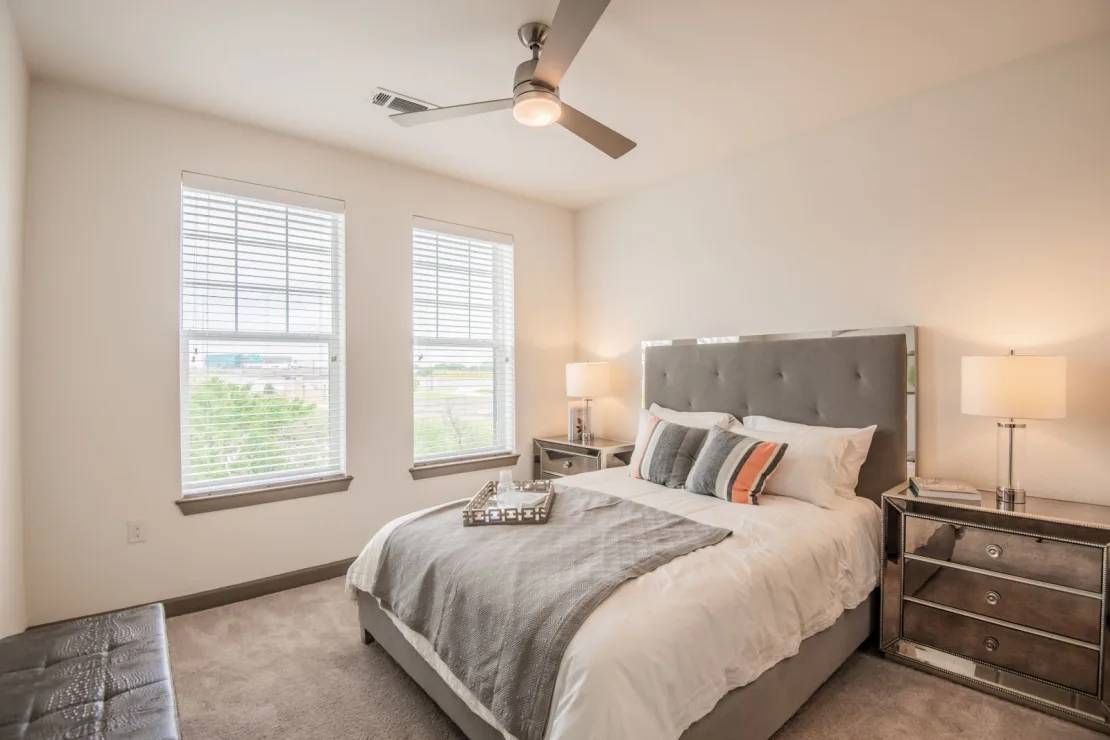 Bright modern bedroom with a gray bed, two windows, bedside lamps, and a ceiling fan