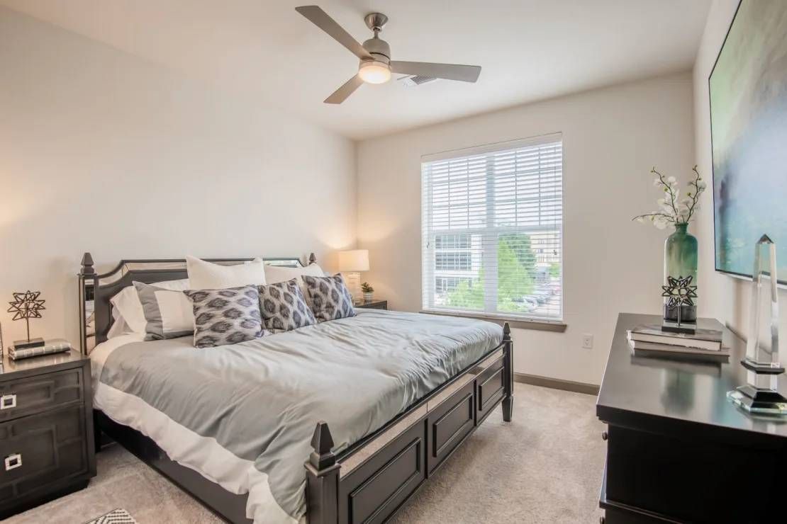 Bright bedroom with a large bed, white walls, window blinds, ceiling fan, and dark dresser.