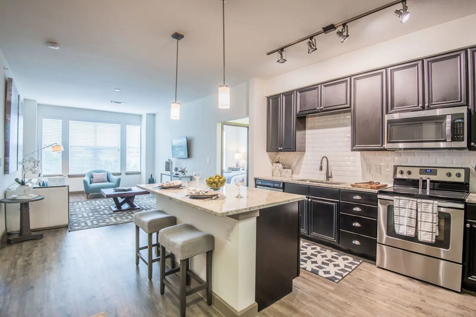 Modern kitchen and living area with island, stools, stainless steel appliances, and dark cabinets.