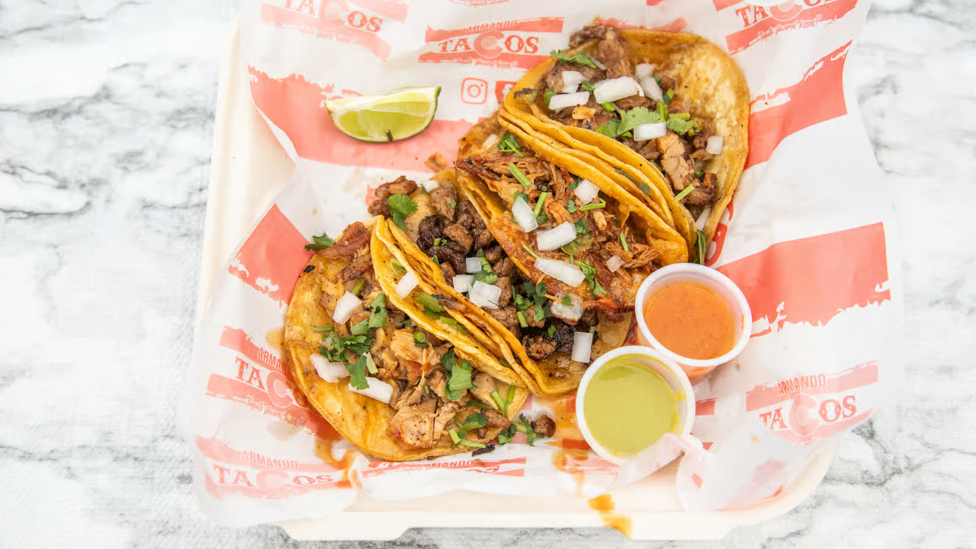 Two tacos on a paper tray with lime wedges and two dipping sauces on a marble table.