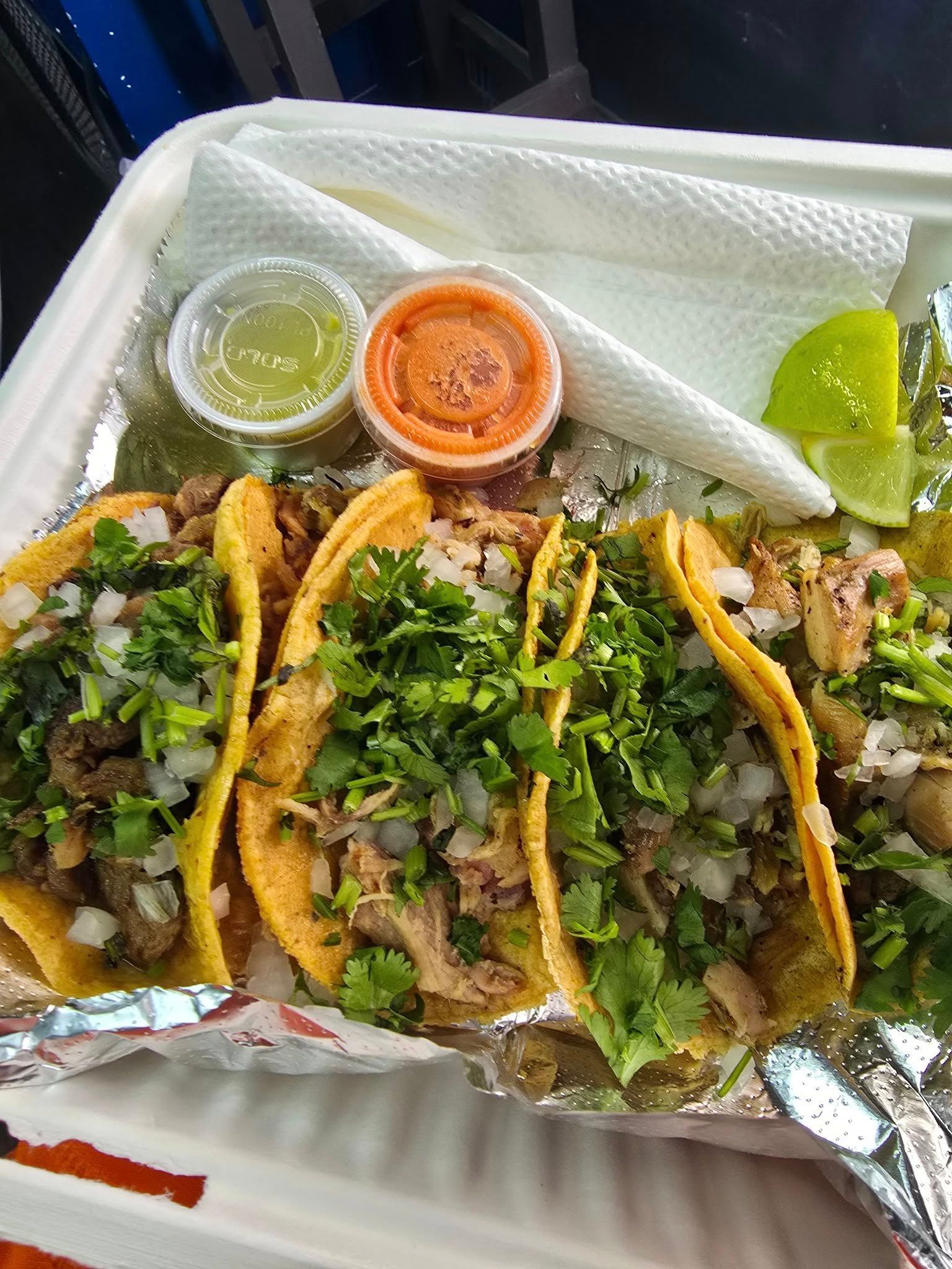 Tray of three tacos with chopped cilantro, lime wedges, and two salsa cups in a foil-lined takeout container