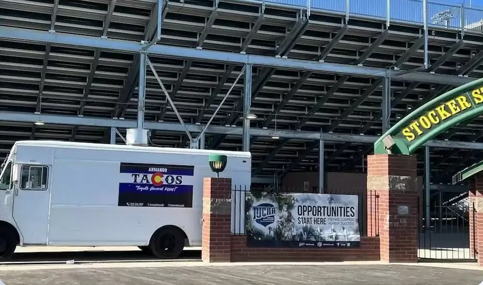 White food truck parked by a brick stadium entrance with a green “STOKKER” sign.