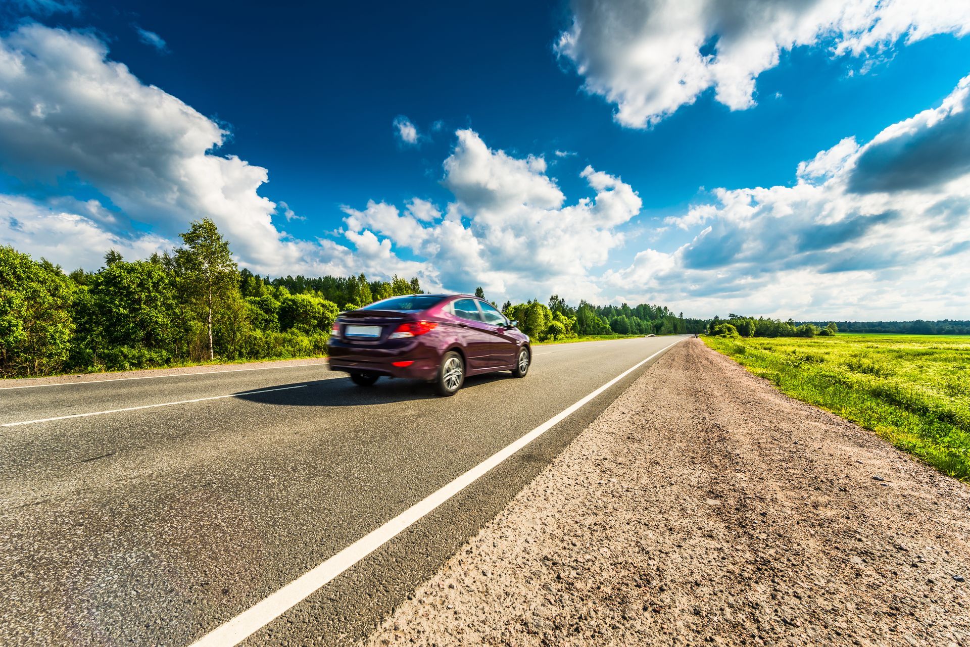 Red car driving on a road with a blue sky and clouds; trees and grass on either side.