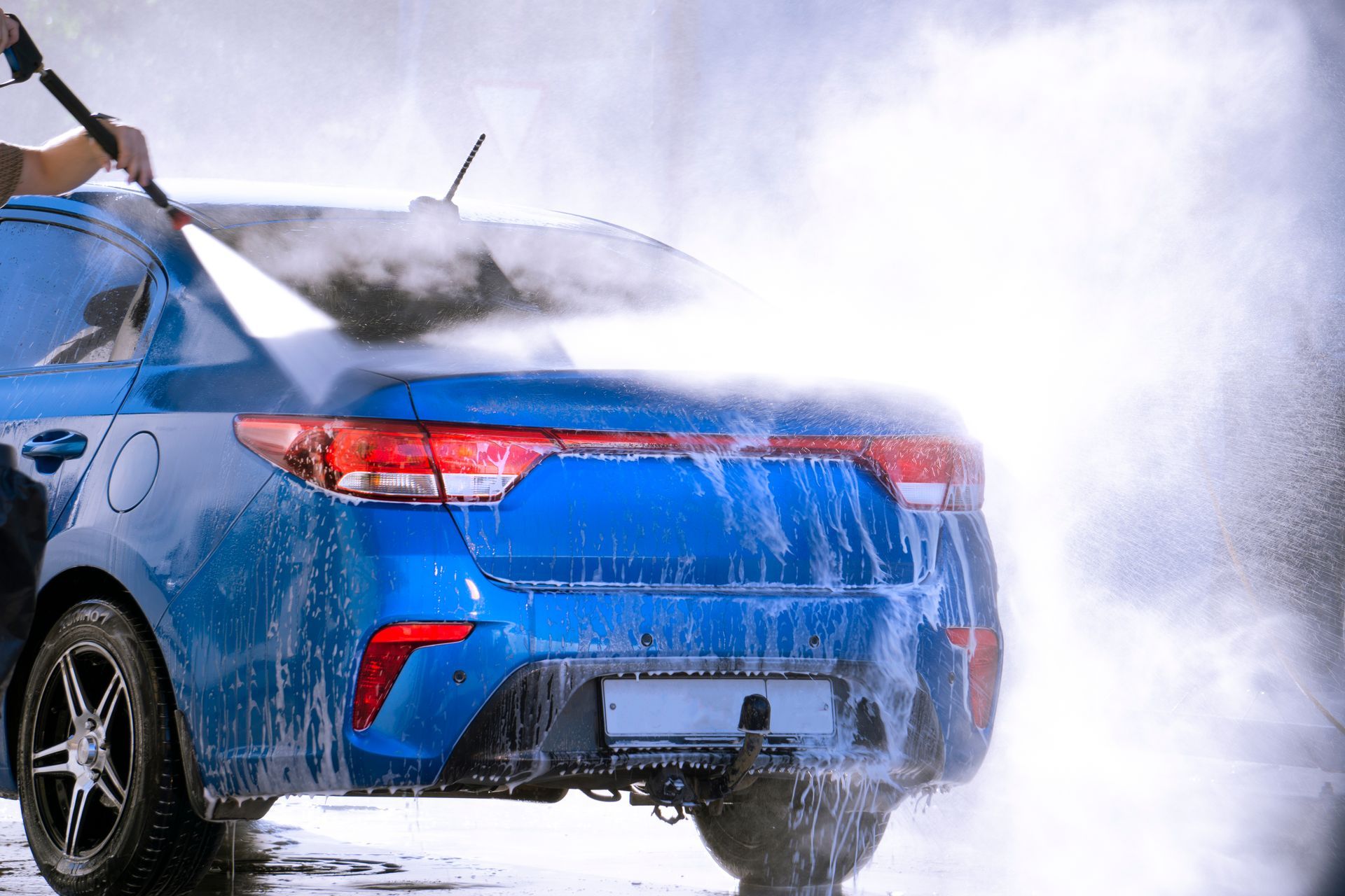 A blue car being washed with a pressure washer, creating a mist.