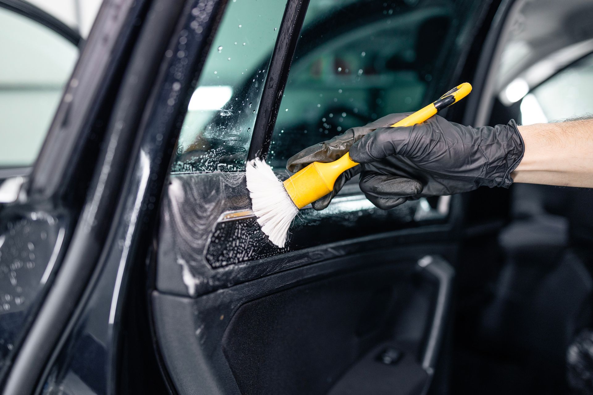 Gloved hand cleaning car door frame with a brush; black car interior, bright yellow brush.