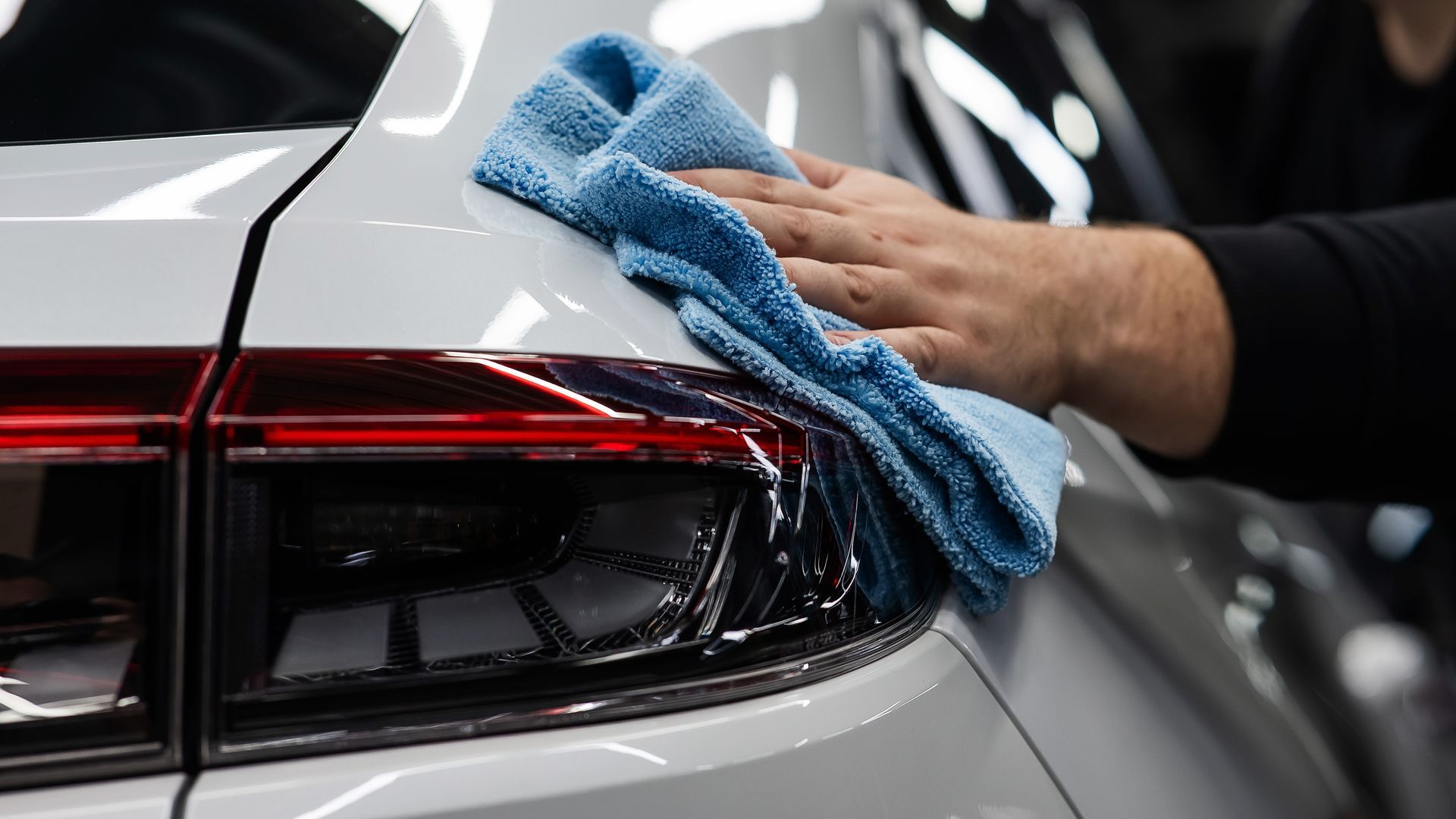 Hand wiping a white car's taillight with a blue microfiber cloth.