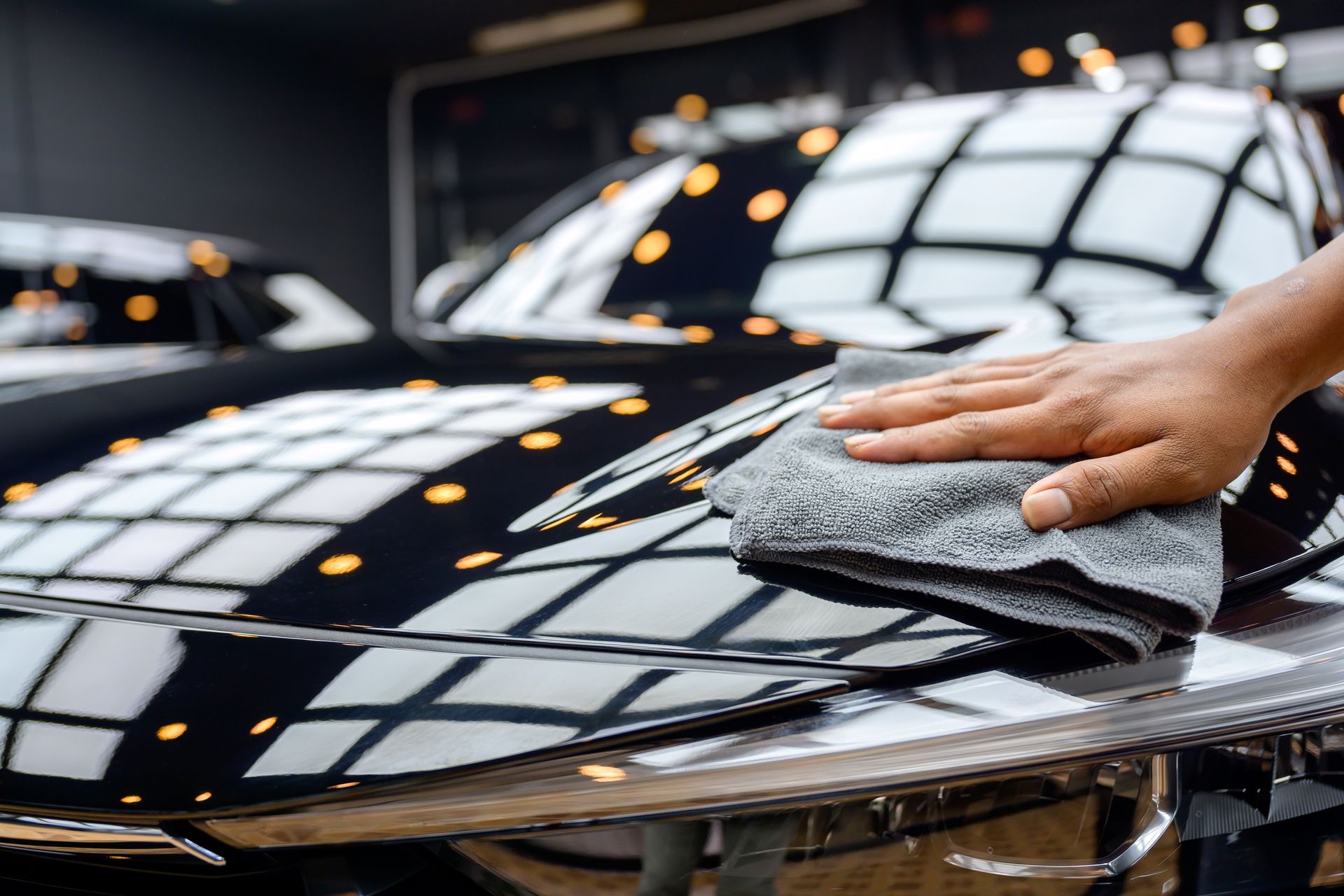 Hand wiping a black car's shiny hood with a gray microfiber cloth. Reflected lights are visible.