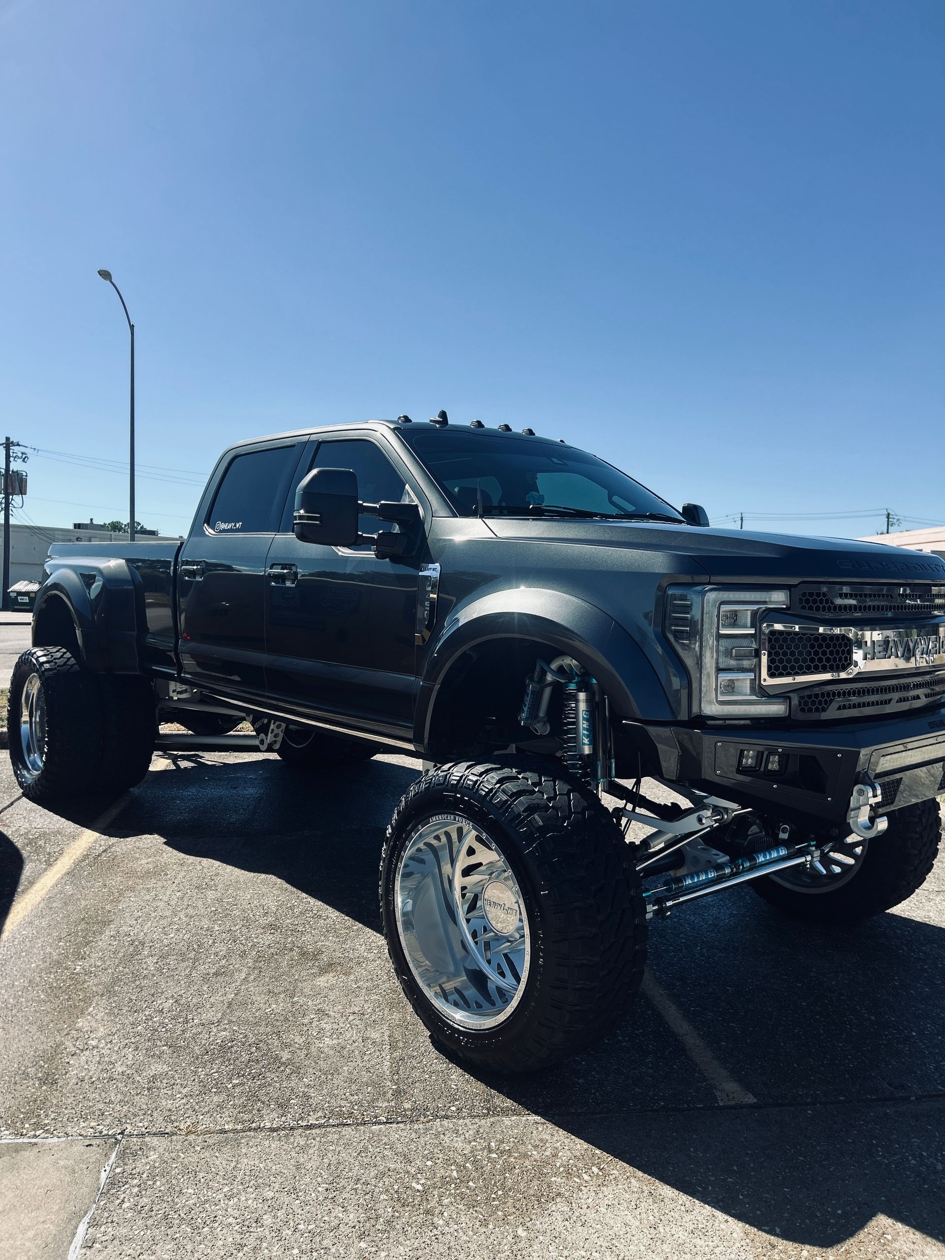 Dark gray, lifted Ford pickup truck with chrome wheels parked outdoors on a sunny day.