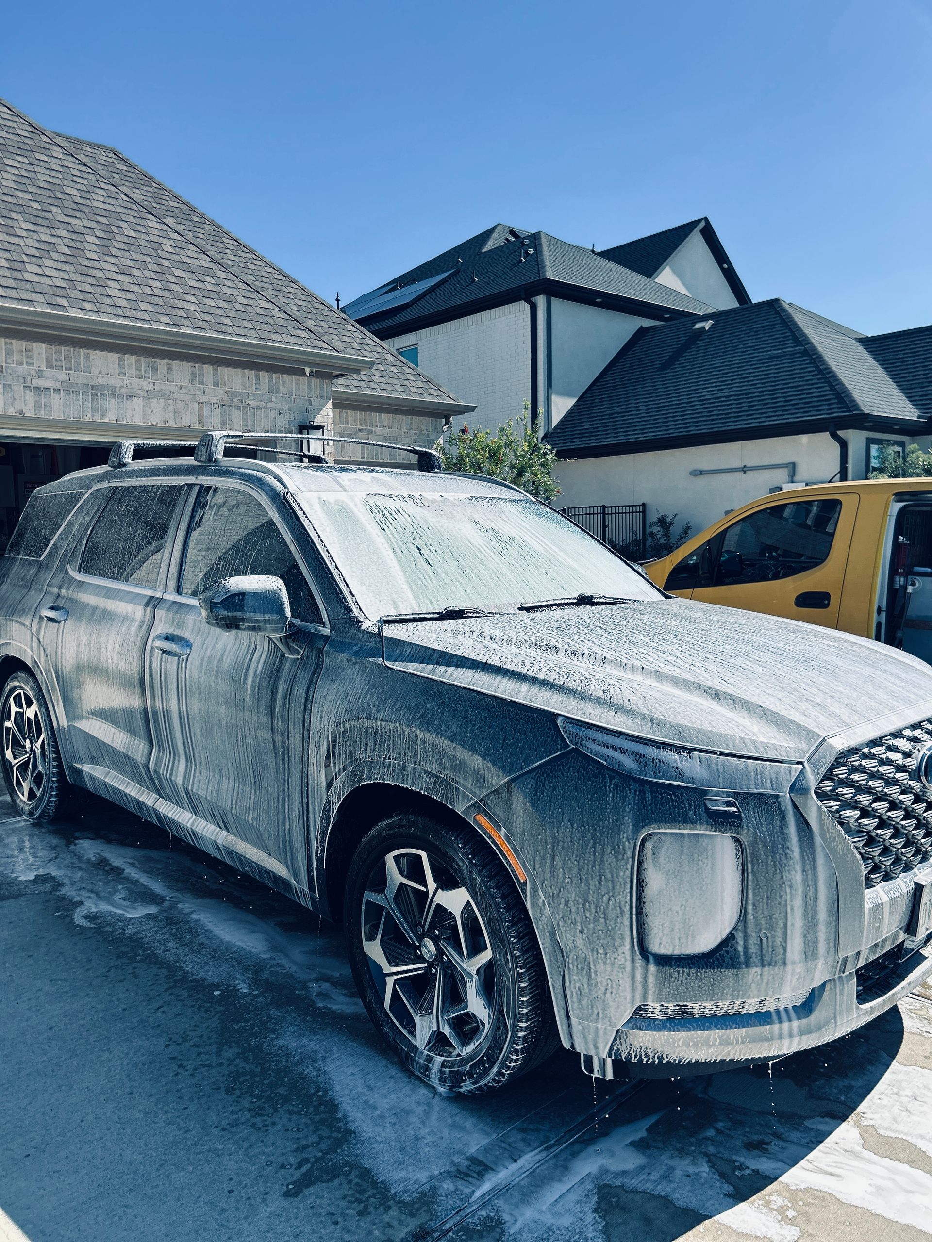 A gray SUV covered in foamy soap suds sits in front of a house, car wash in progress.