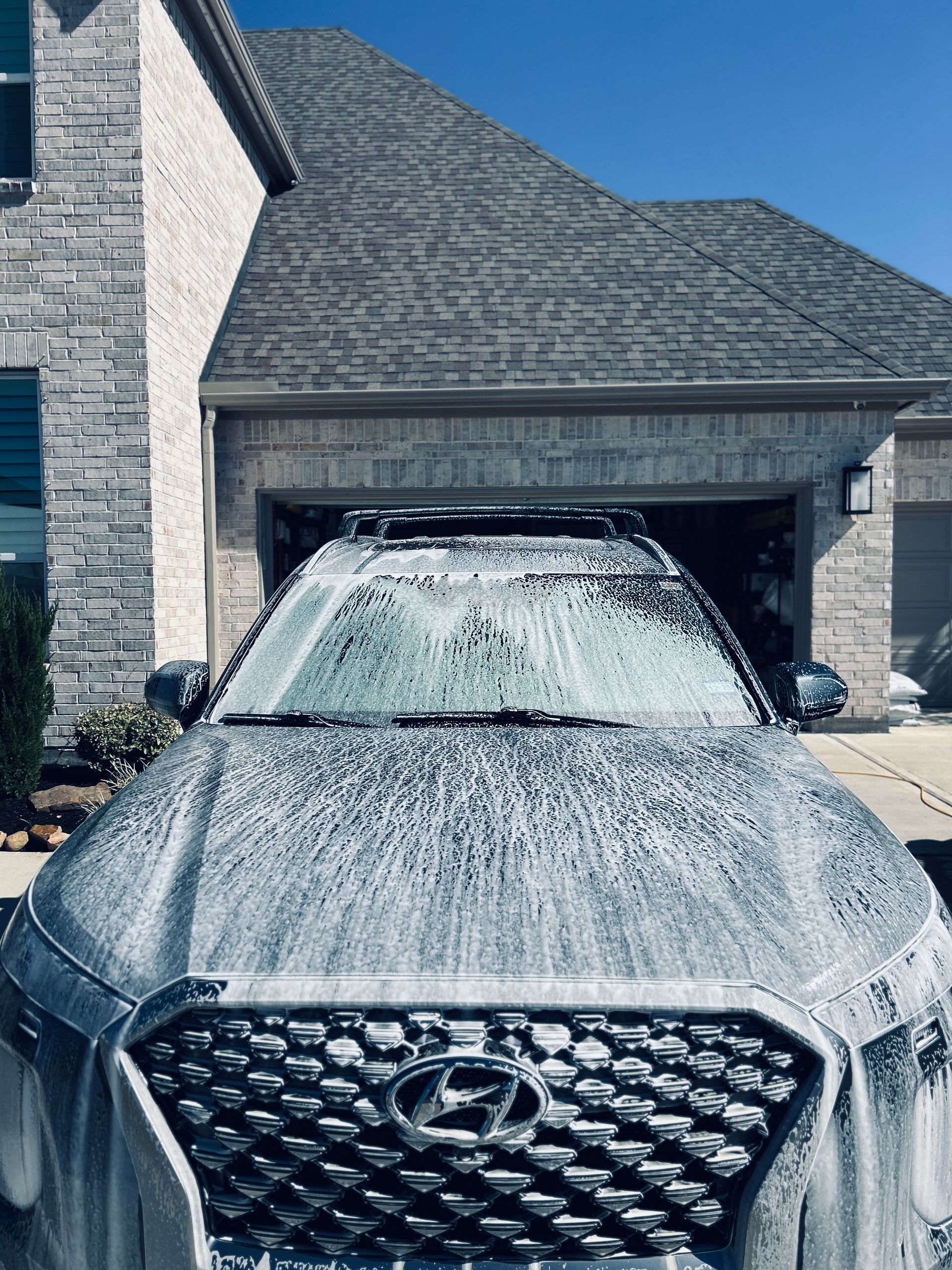 Car covered in white soap suds in front of a house, being washed on a sunny day.