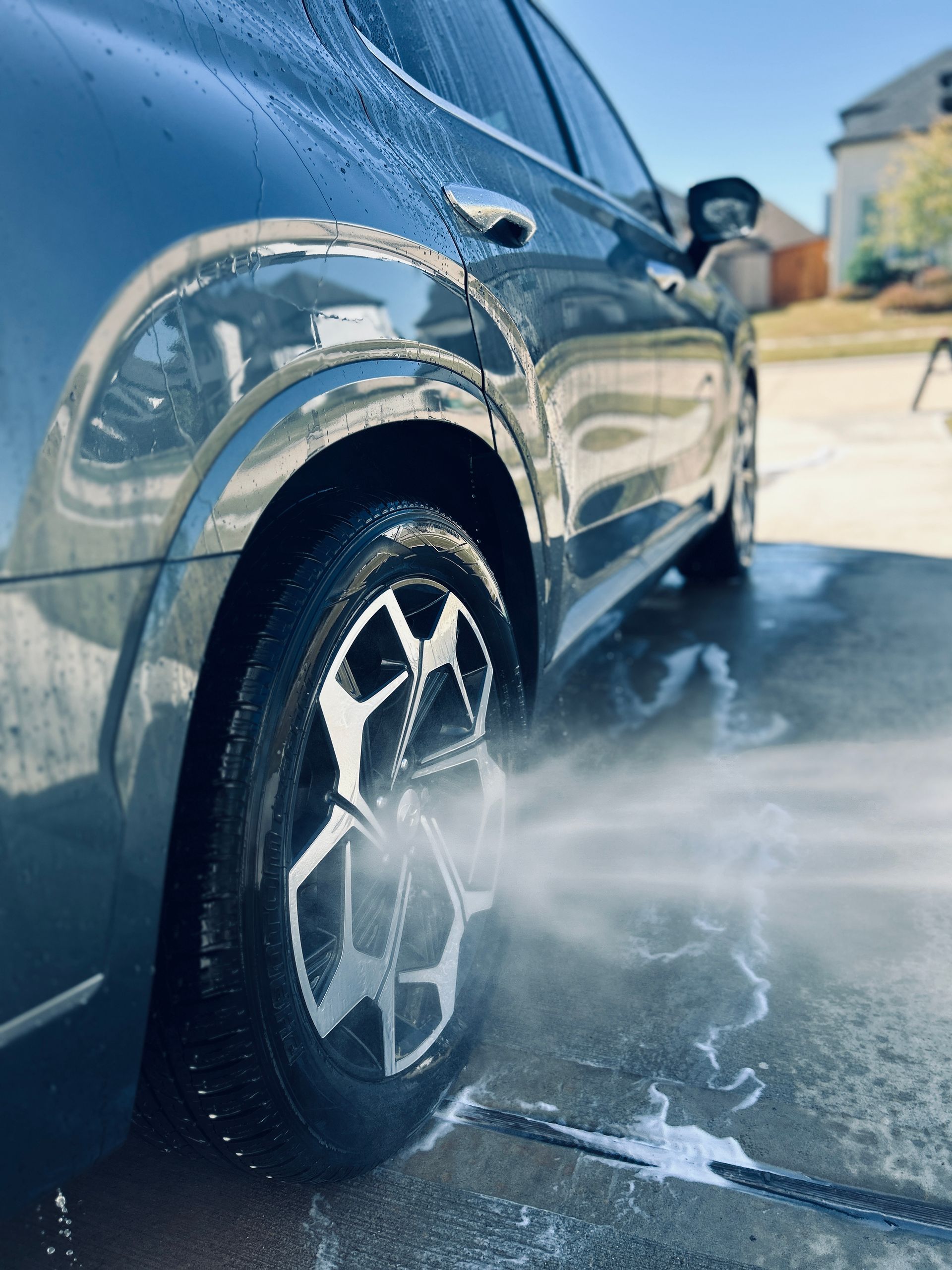 A dark gray car being washed, water spraying from a hose onto a tire.