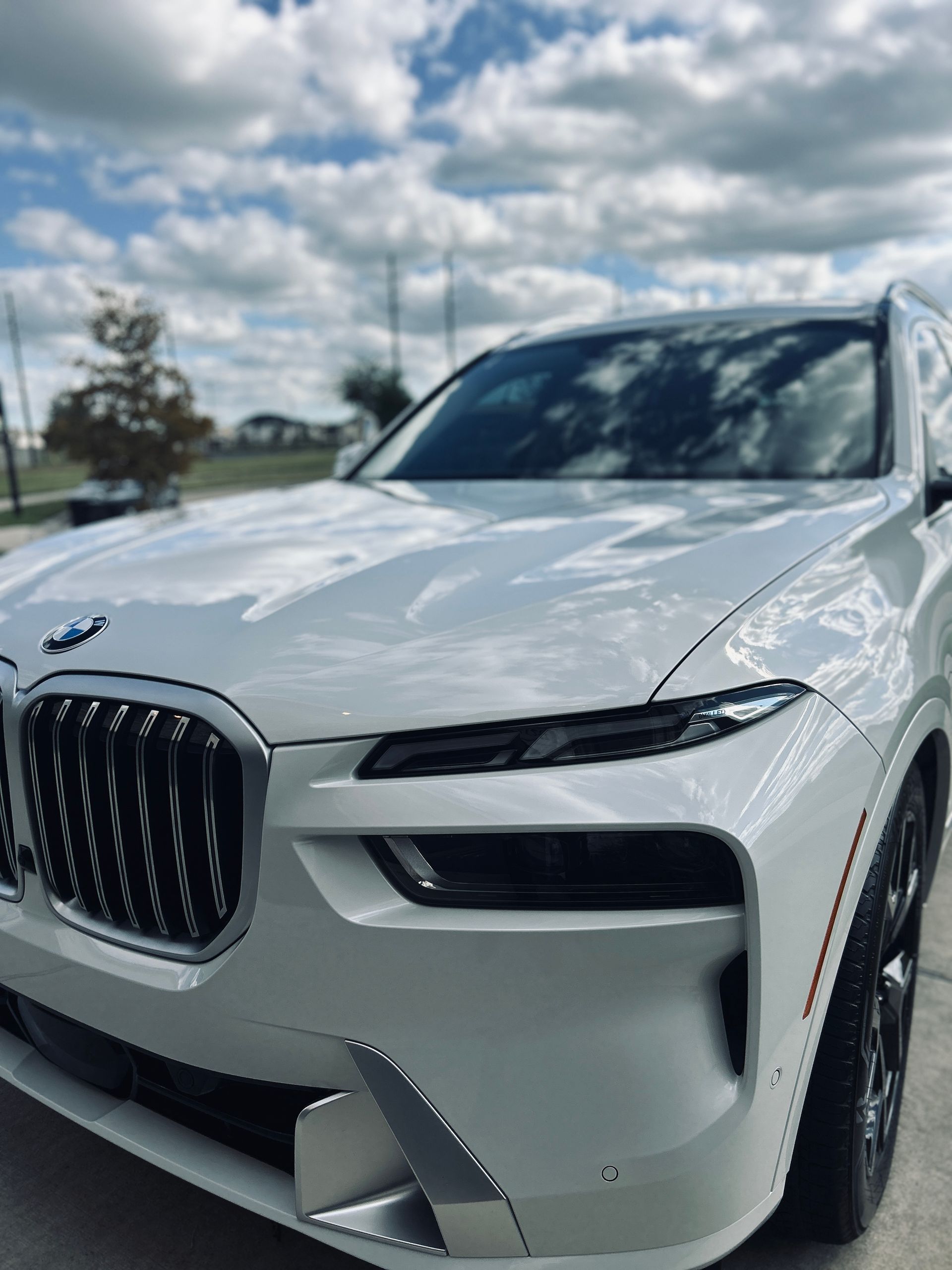 White BMW X7 SUV parked outdoors, shiny finish, against a blue sky with some clouds.