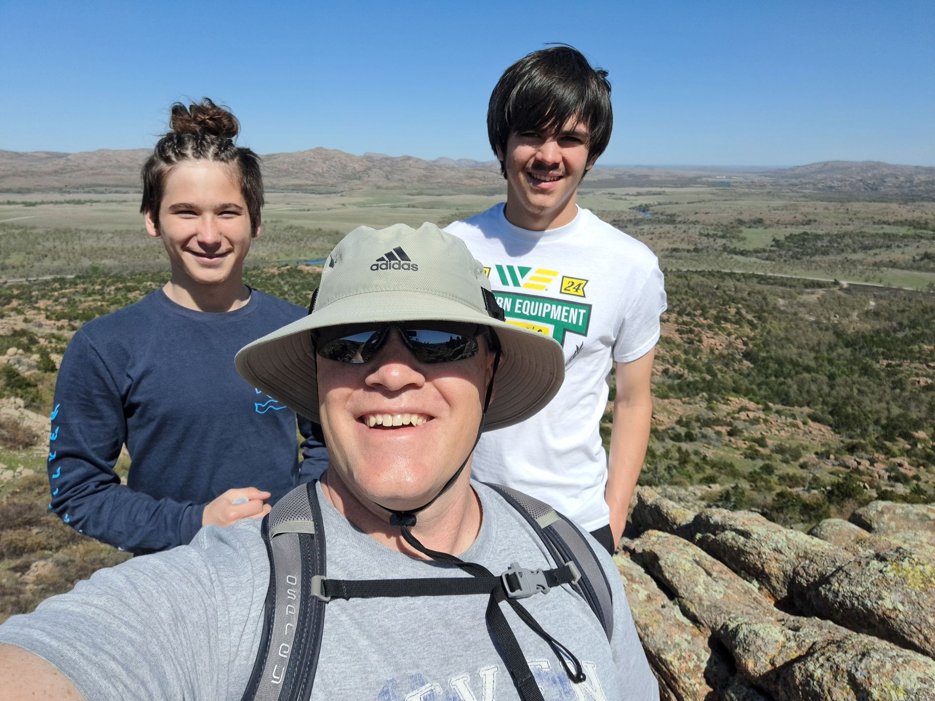 Three people pose for a selfie on a rocky mountain summit against a clear blue sky.