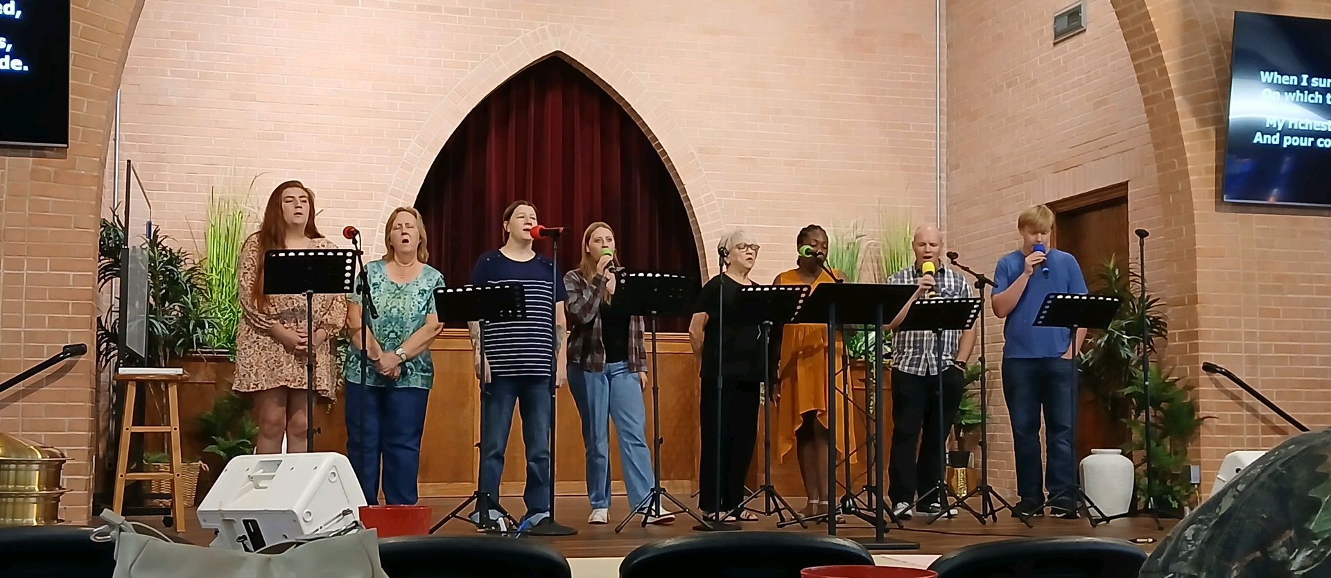 A group of eight people stands in a line on a church stage, singing into microphones behind music stands.