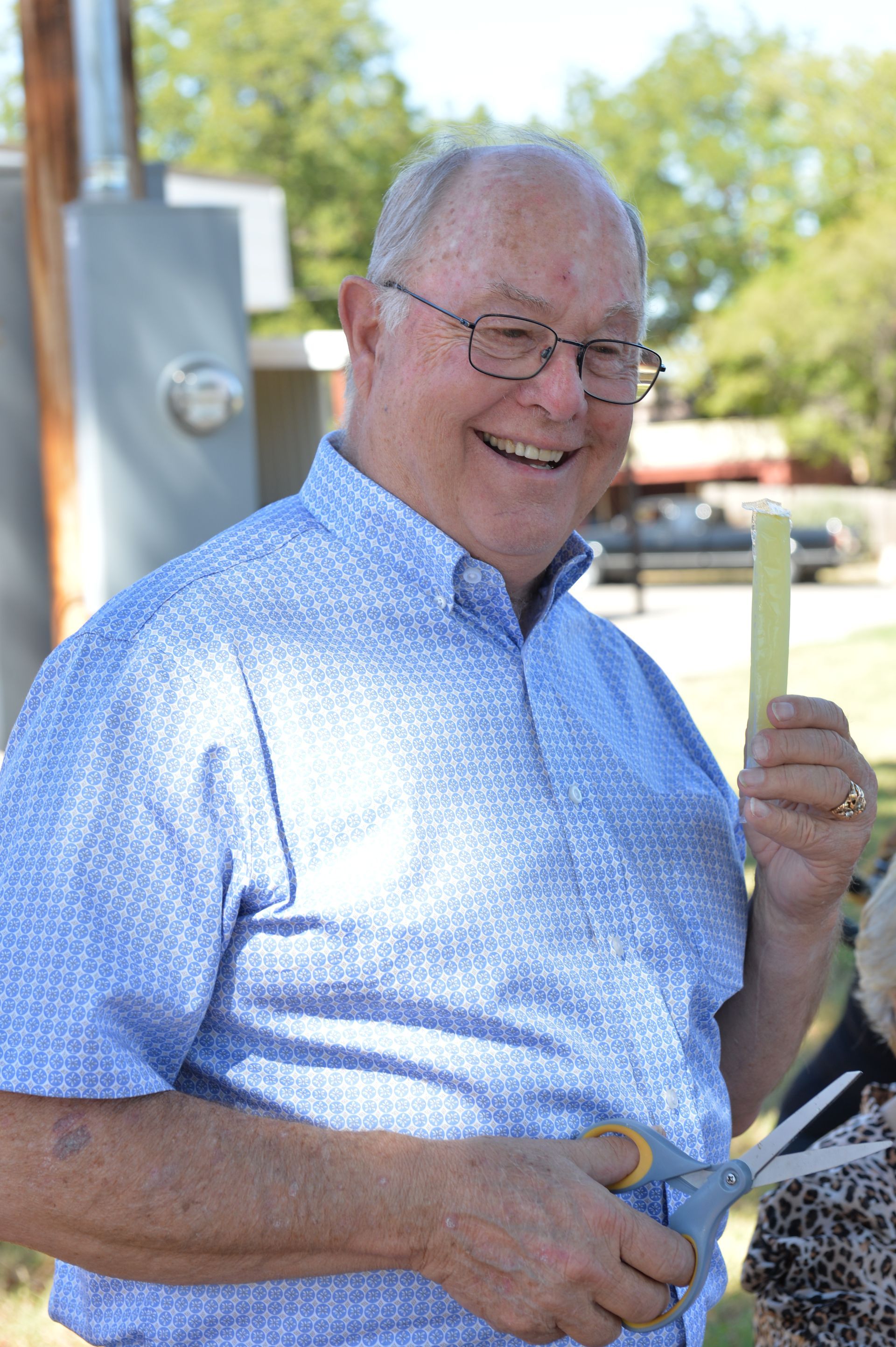 Smiling person in a blue patterned shirt holding scissors and a small yellow ribbon strip outdoors.