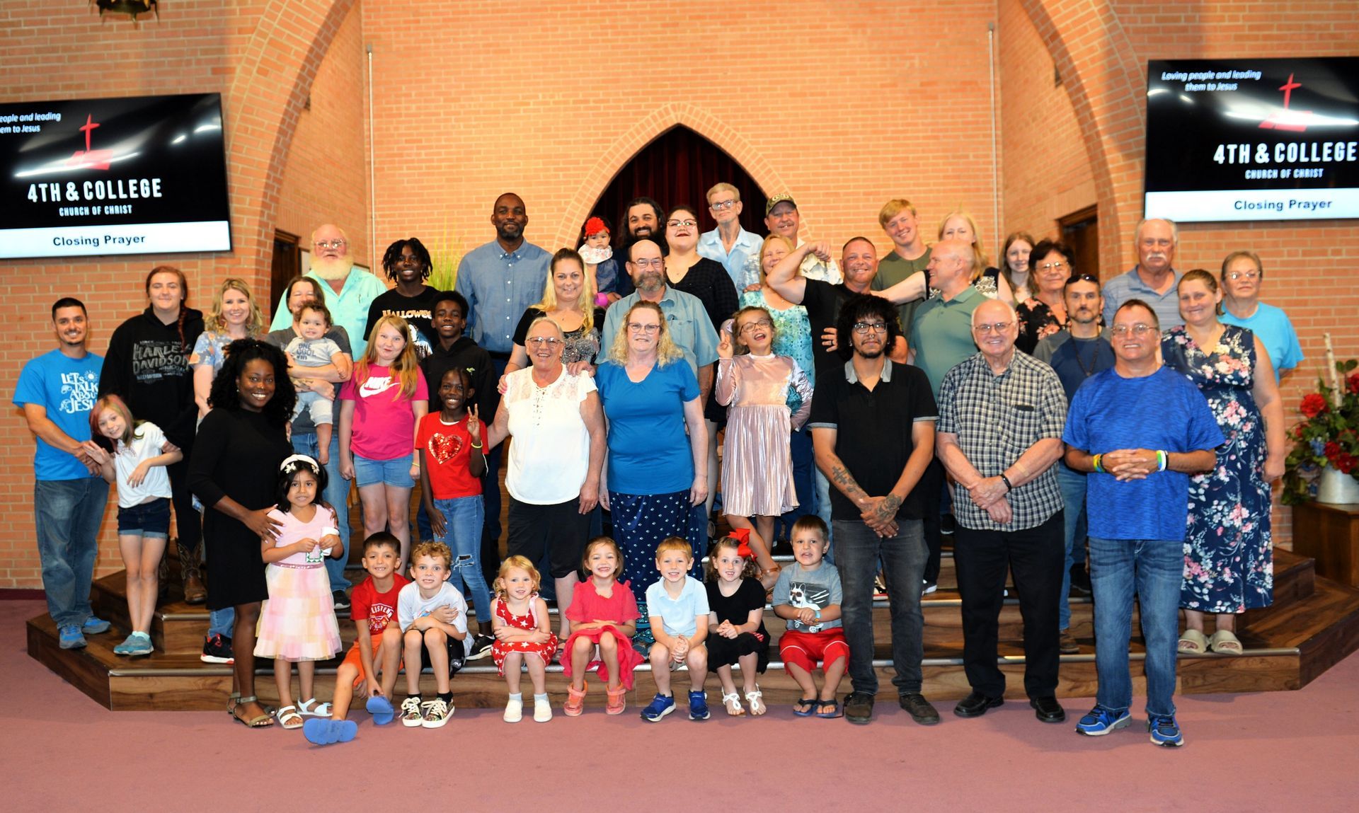 A large group of people poses on a stage in front of a brick archway and two large screens in a church setting.