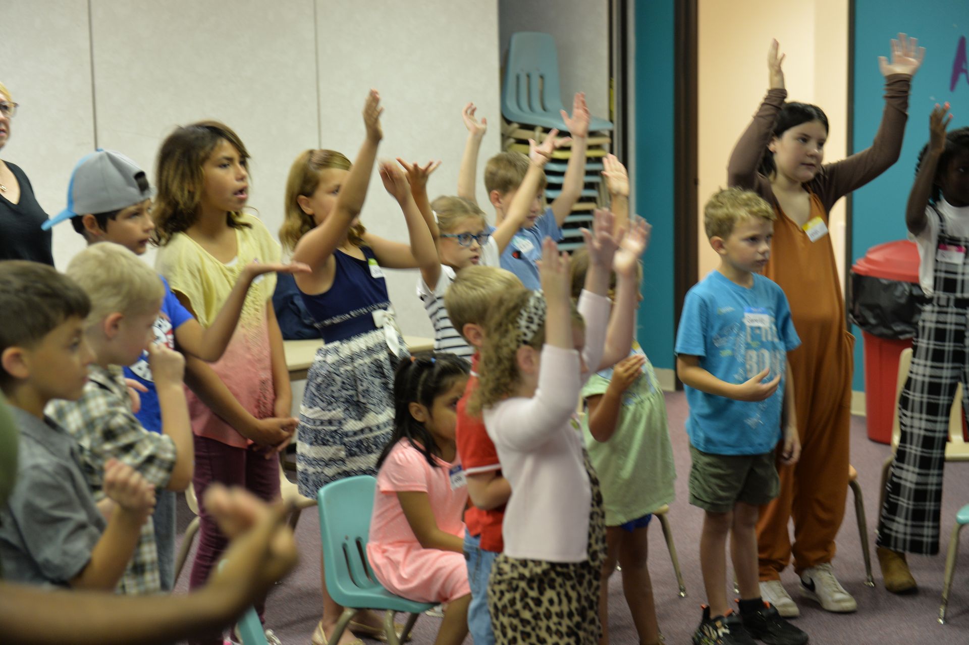 A group of children in an indoor room standing and participating in an activity with hands raised.