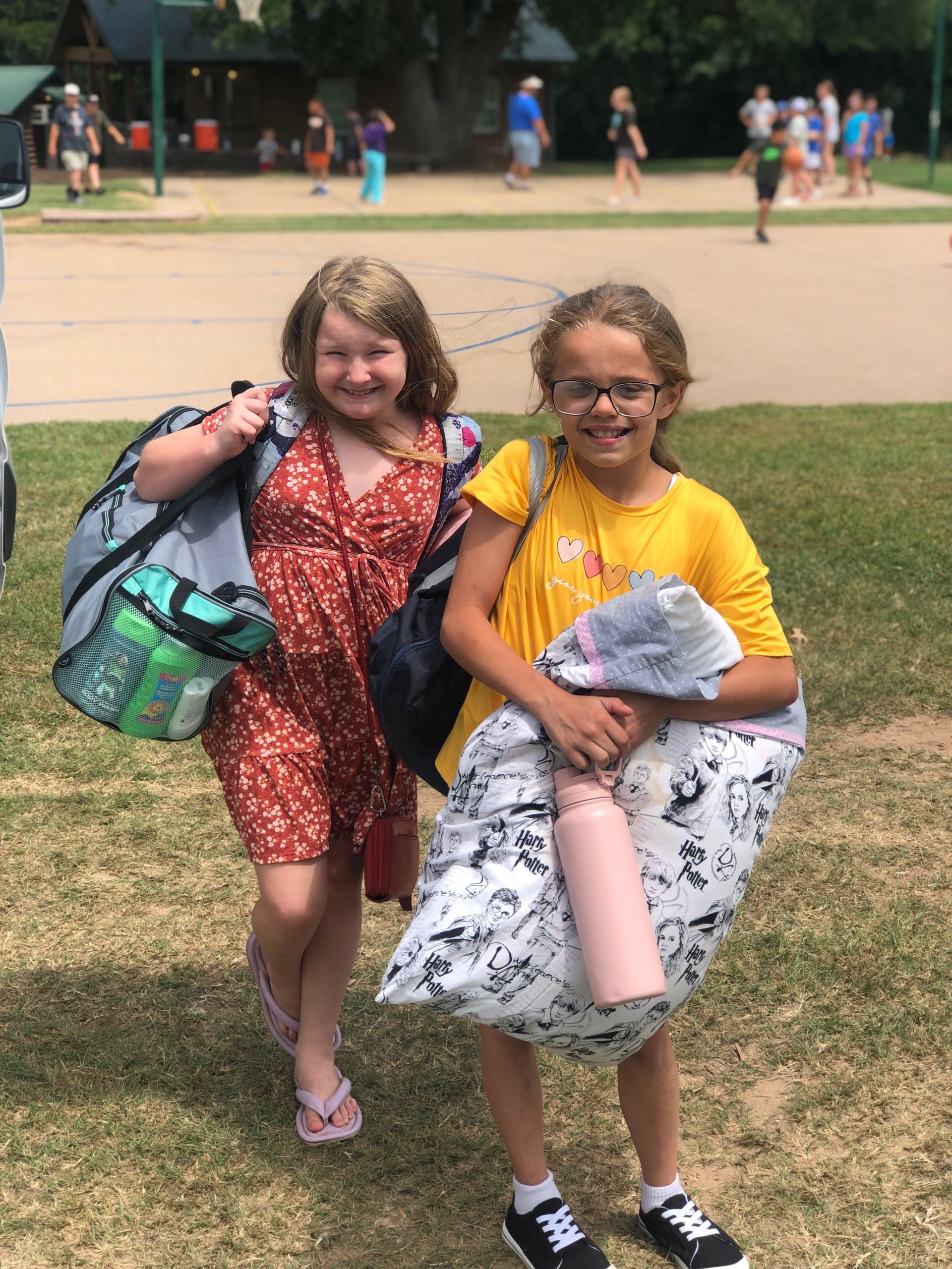 Two children stand outdoors smiling, holding backpacks and gear, with a camp or park setting in the background.
