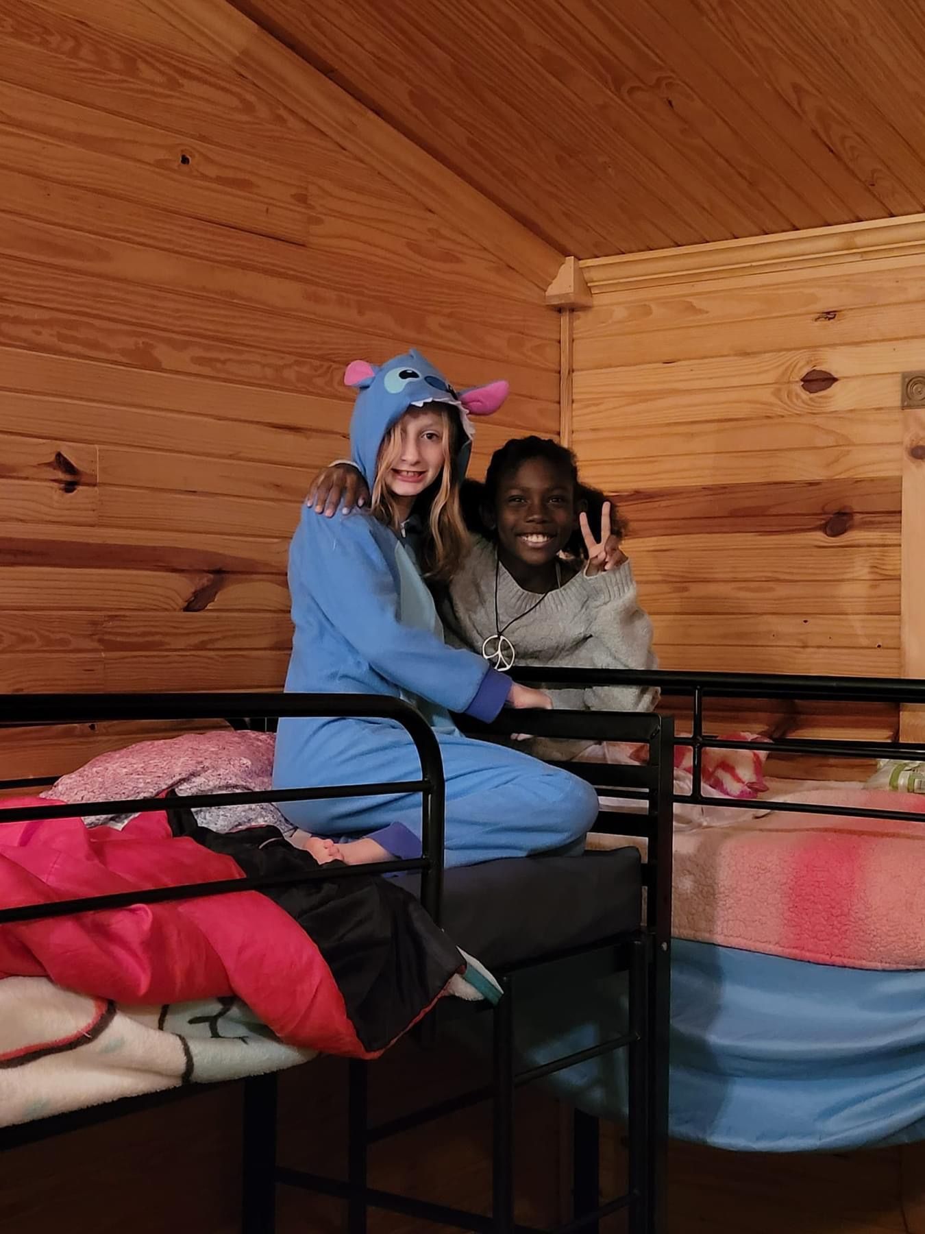Two people sitting on bunk beds in a room with wood-paneled walls; one wears a blue character onesie and the other smiles.