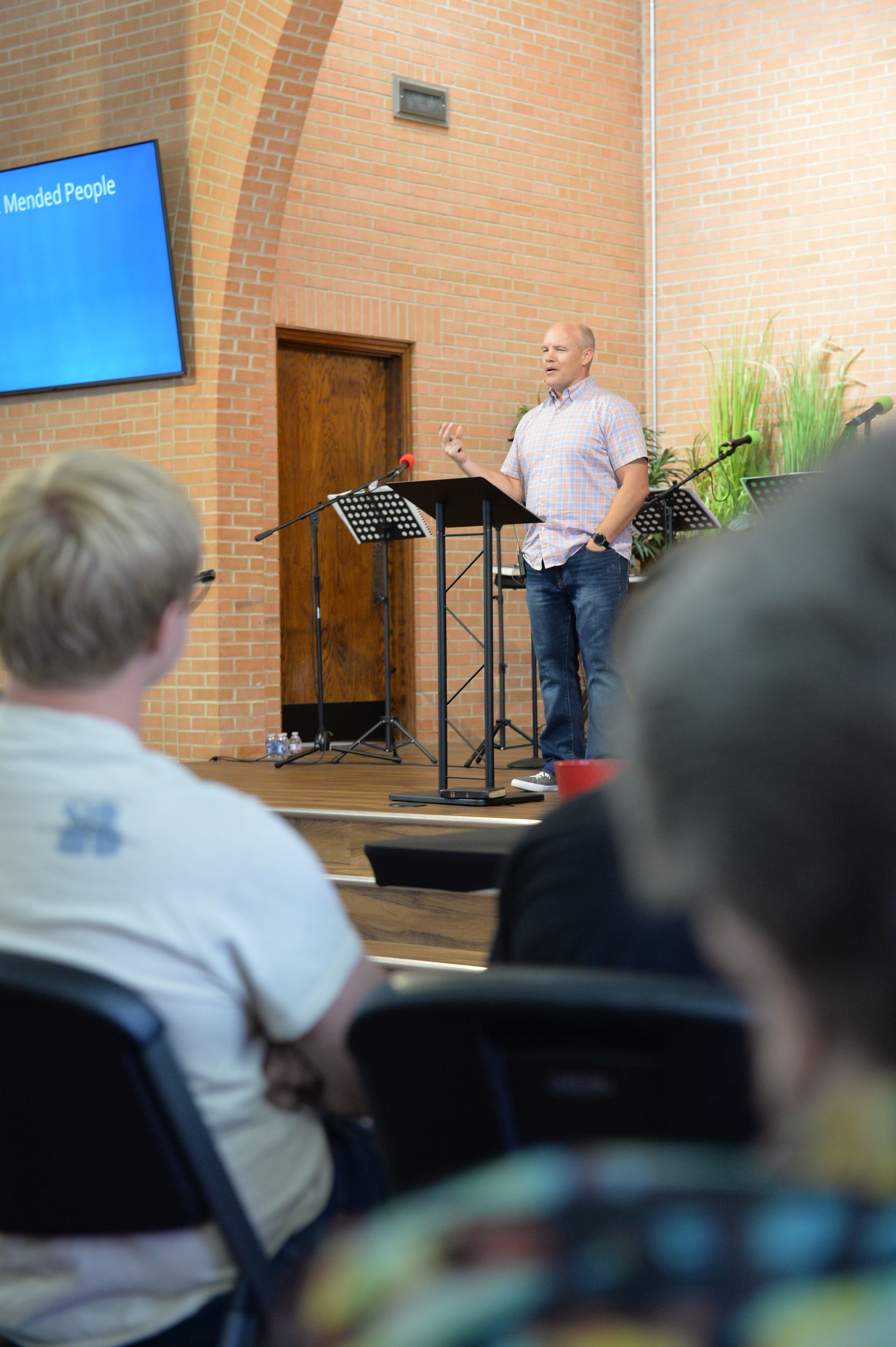 A speaker stands at a podium in a brick-walled auditorium, addressing an audience from a stage.