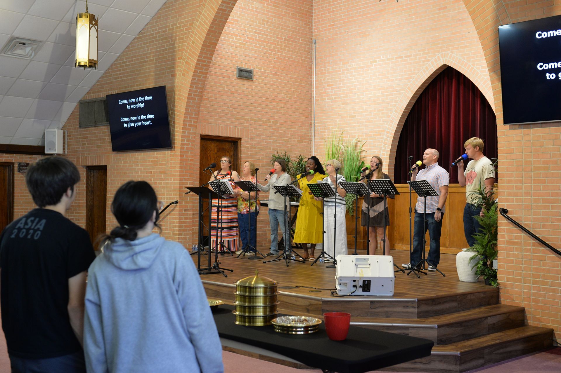 A group of people standing on a stage in a church, singing into microphones with music stands in front of them.
