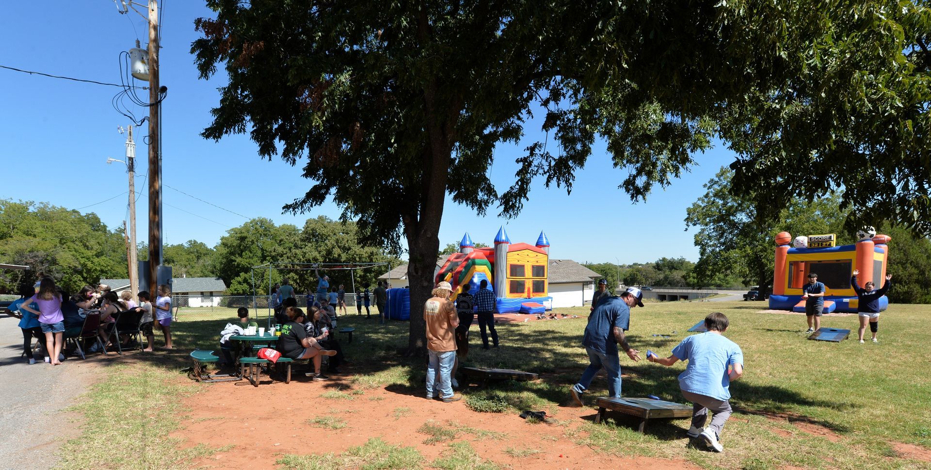 People gathering in a park with bouncy castles and yard games under a large tree on a sunny day.