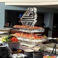 Buffet table with stacked plates, trays of salad and appetizers, and a tiered clear centerpiece in a dining hall