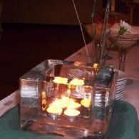 Small glowing candle in a glass cube on a table with a green napkin and glassware nearby