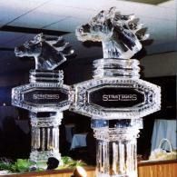 Two silver horse-head ice sculpted trophies with black nameplates on a table indoors under a ceiling.