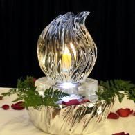 Decorative glass sculpture with a glowing candle, surrounded by greenery and red rose petals on a table